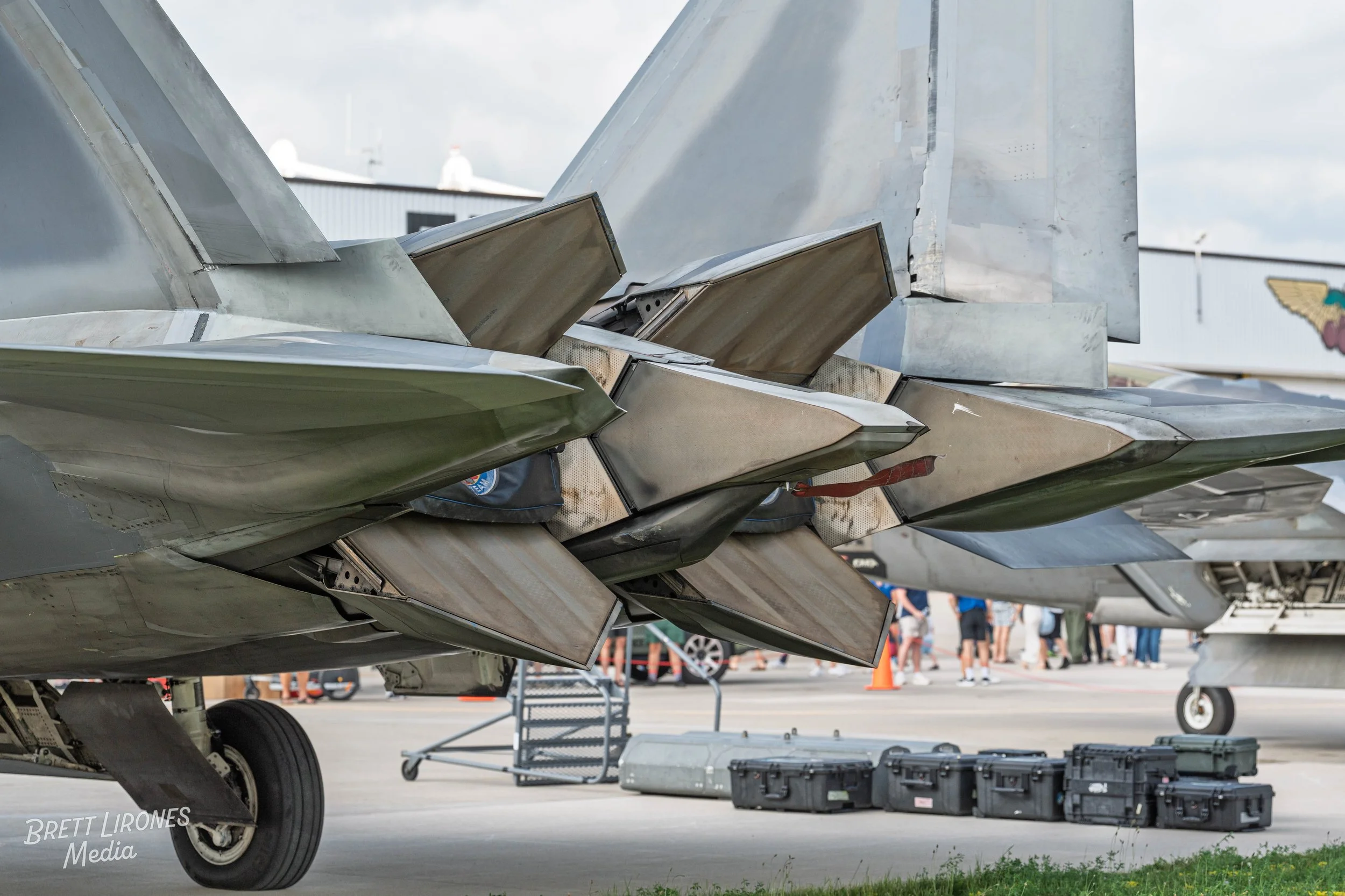 Close-up of the back of a fighter jet, showing the tail fins and exhaust nozzles, with people and equipment in the background.
