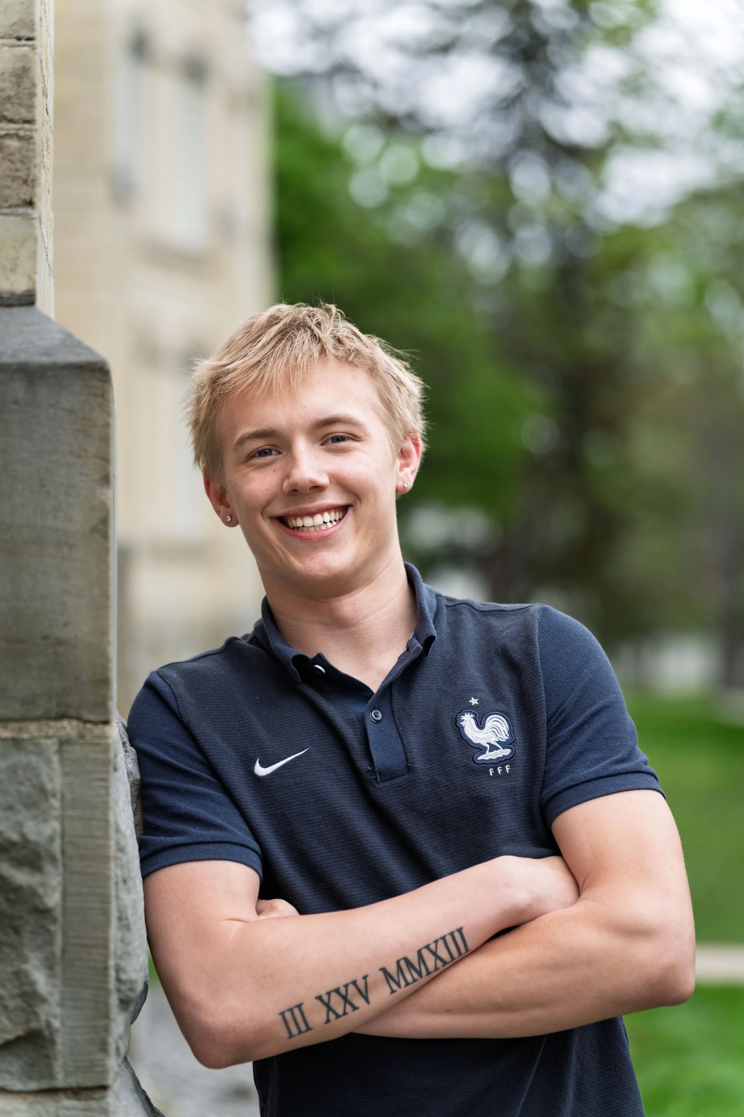 Young man with blonde hair smiling, wearing a navy blue France national football team shirt, standing outdoors near a stone wall, with trees in the background.