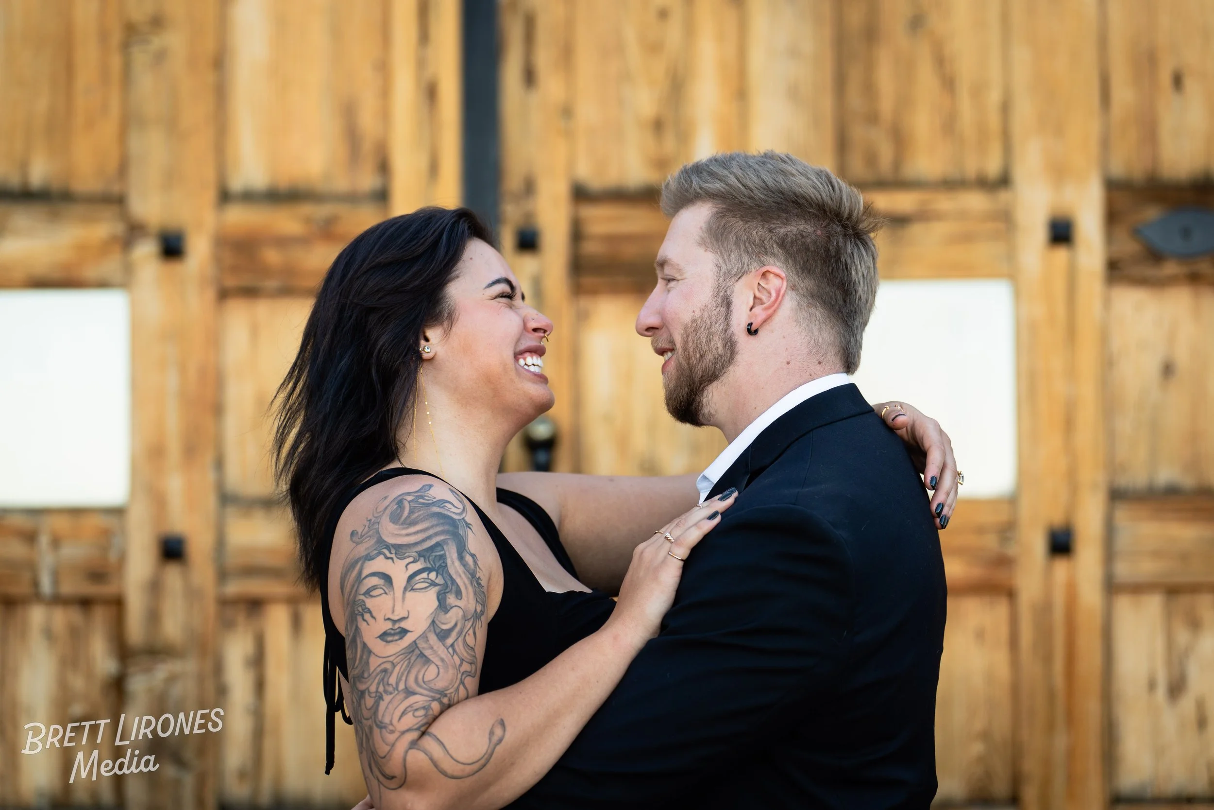 A smiling woman with dark hair and tattoos on her arm embracing a man in a black suit against a wooden wall background.