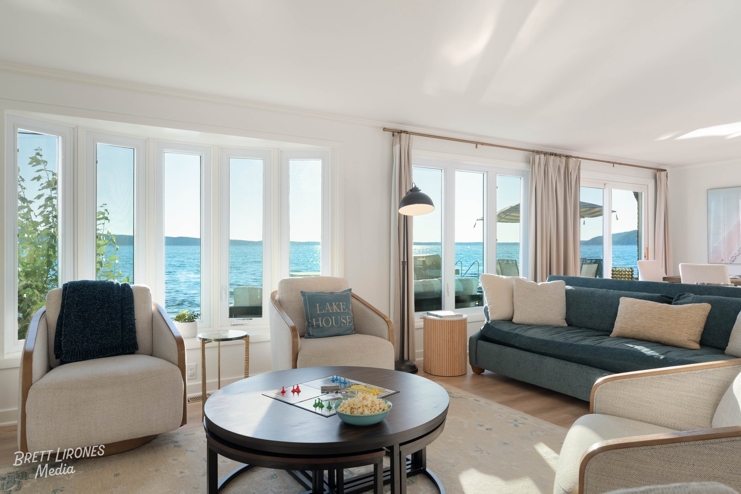 Living room with ocean view, beige and dark blue couches, coffee table with popcorn and game pieces, large windows showing lake and outdoor patio