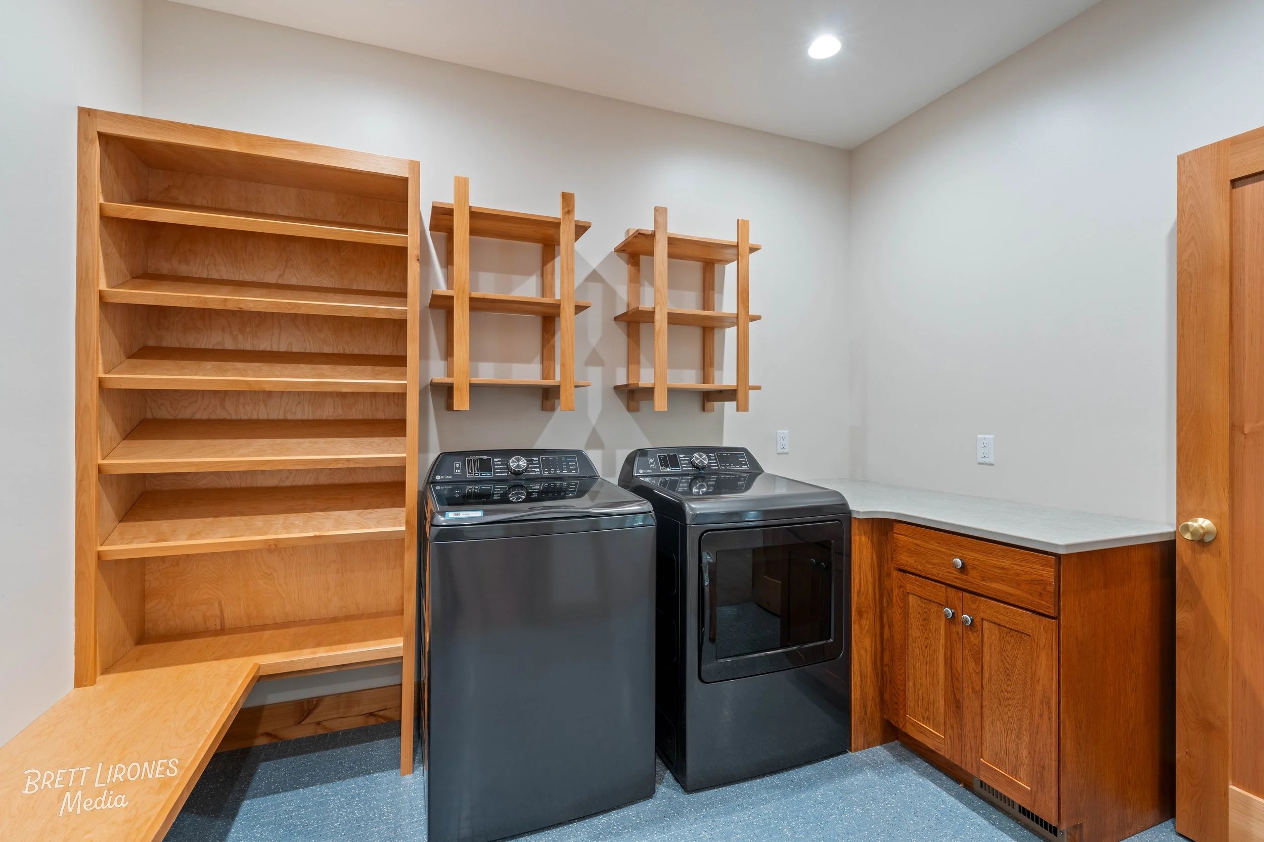 A laundry room with a black washer and dryer, wooden shelves and cabinets, and a white countertop on the right side.
