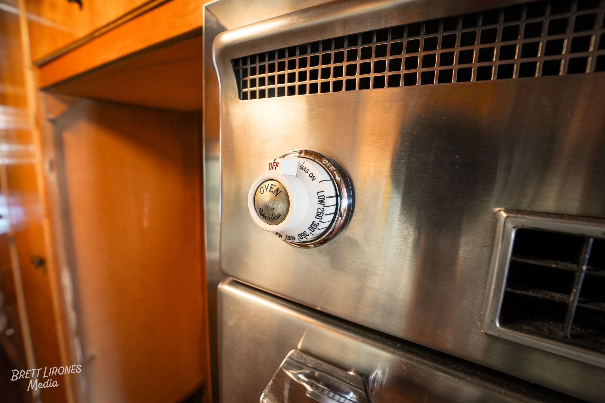 Close-up of a stainless steel gas stove burner control knob set to 'Oven' position, with a temperature range from 150 to 500 degrees, in a kitchen with wooden cabinetry.