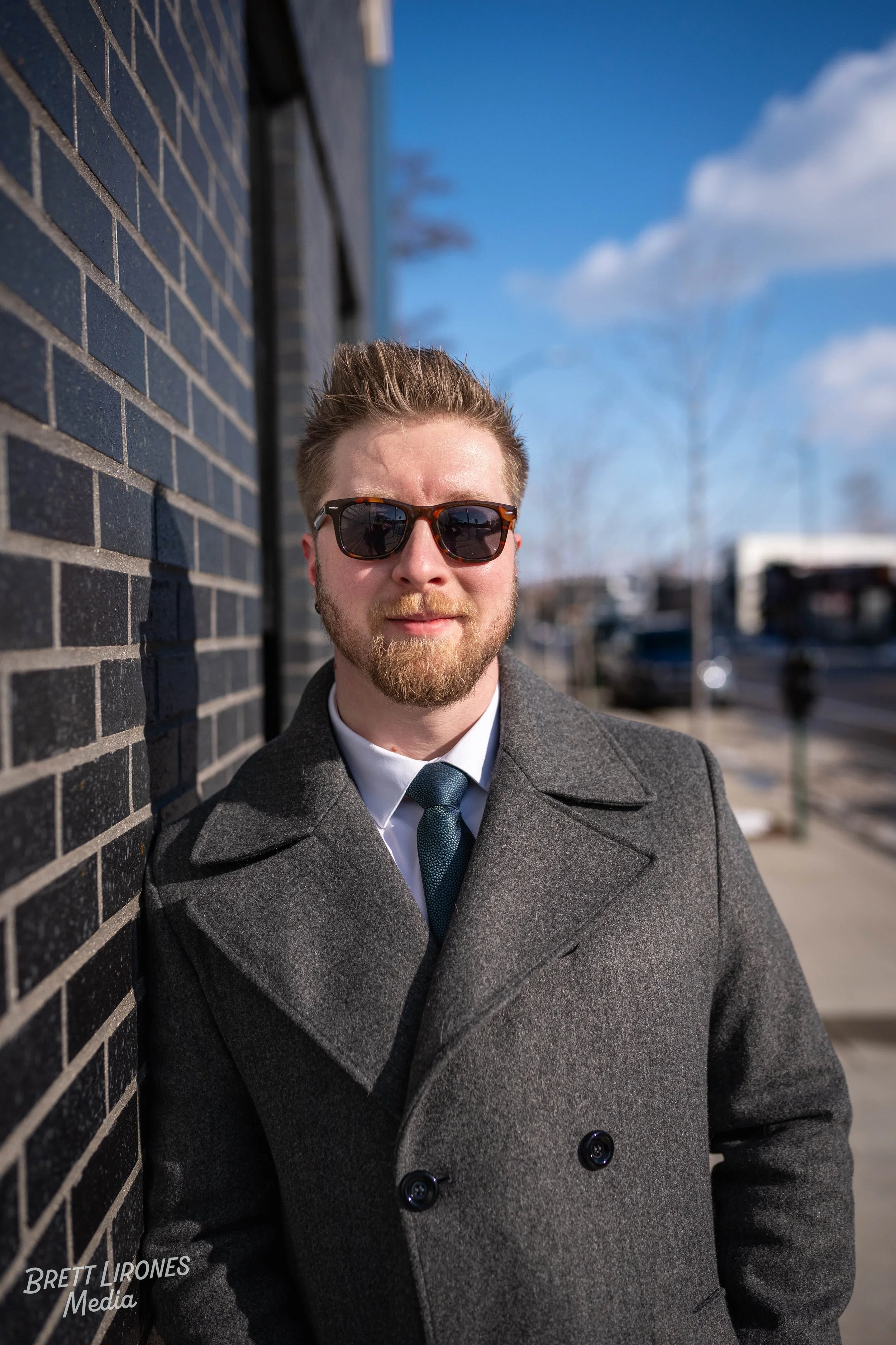 A man with light brown hair, a beard, and sunglasses leaning against a brick wall on a sunny day, wearing a gray coat, white shirt, and a teal tie, with a blurred city street in the background.