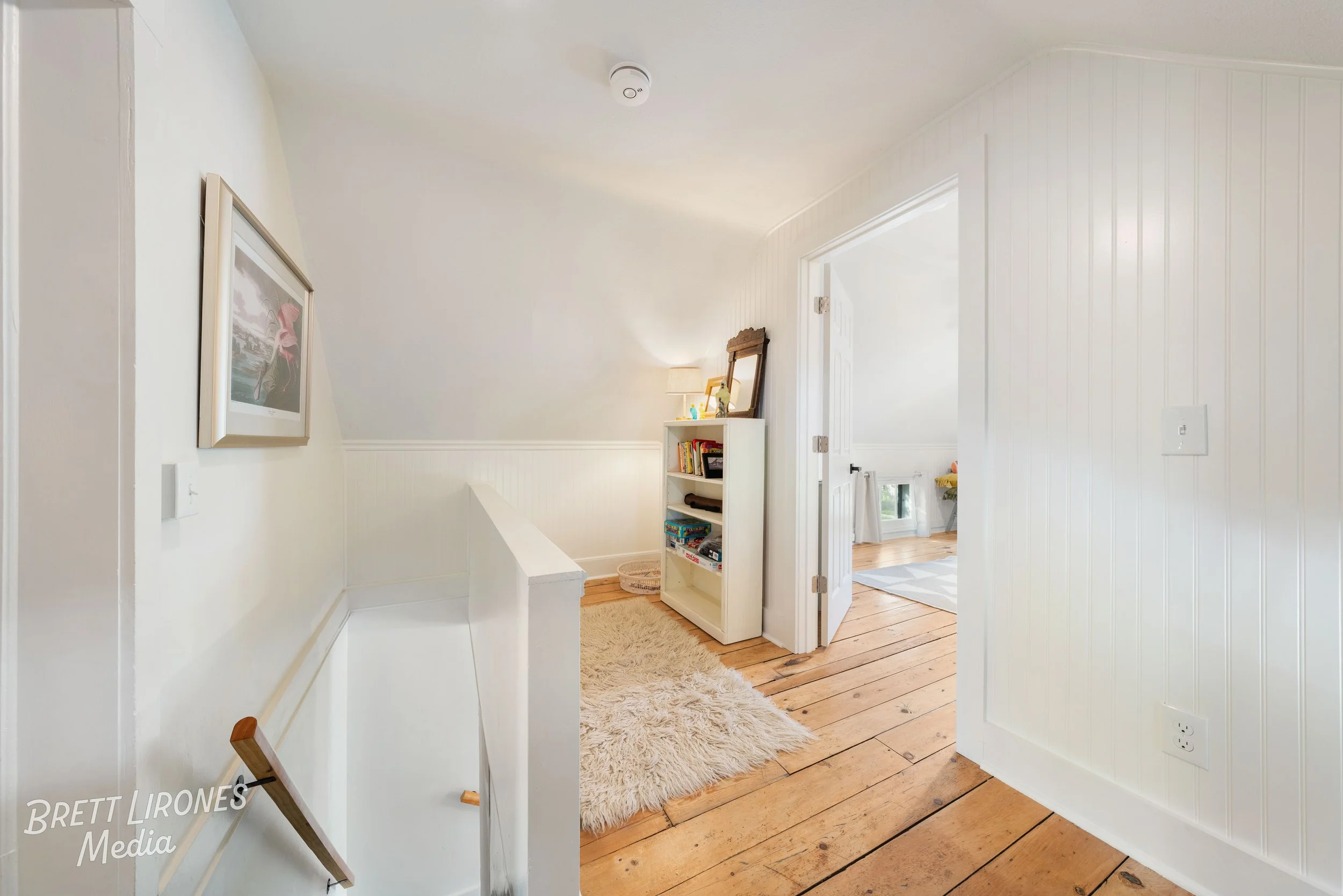 Foyer with white walls, wooden flooring, a fluffy cream-colored rug, a small white bookshelf, and a doorway leading to another room with windows and decor.