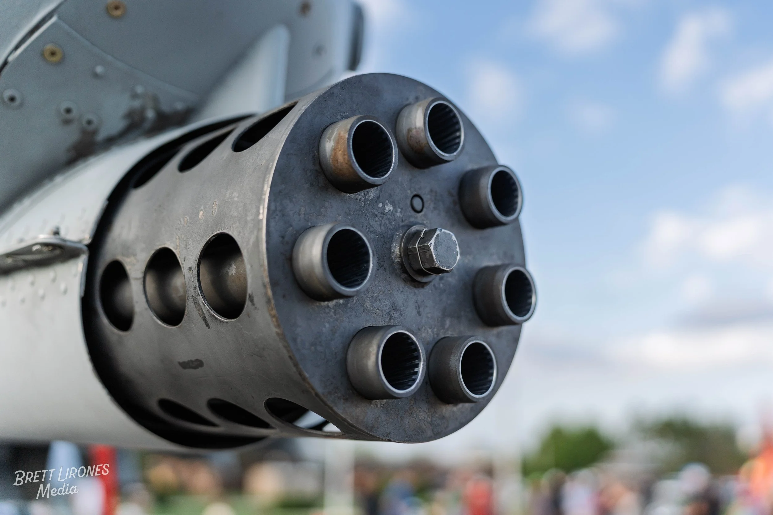 Close-up of a machine gun barrel with multiple vents, part of a military aircraft or vehicle, against a partly cloudy sky background.
