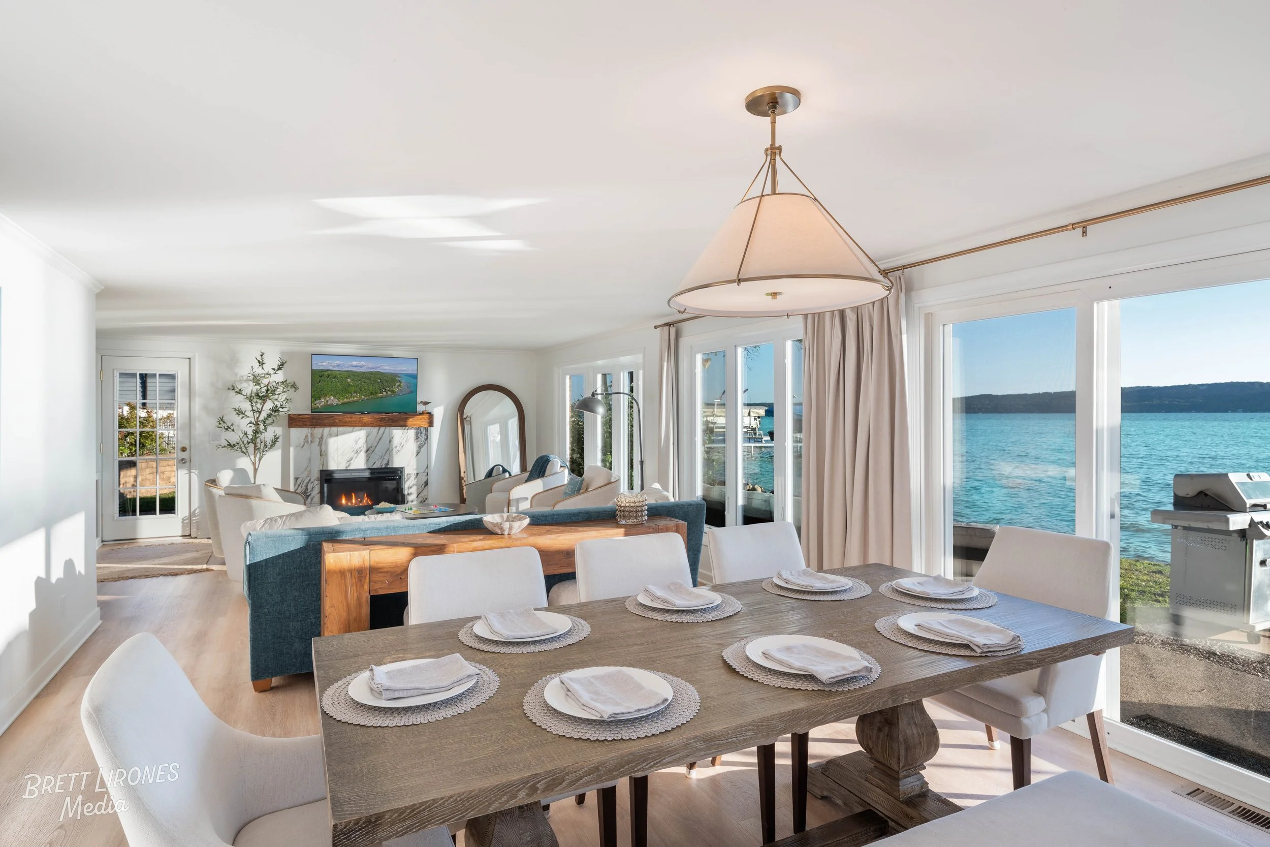 Open-concept living and dining area with large windows showing a waterfront view, a dining table set with white plates and napkins, a fireplace, chairs, and a TV mounted above the fireplace.