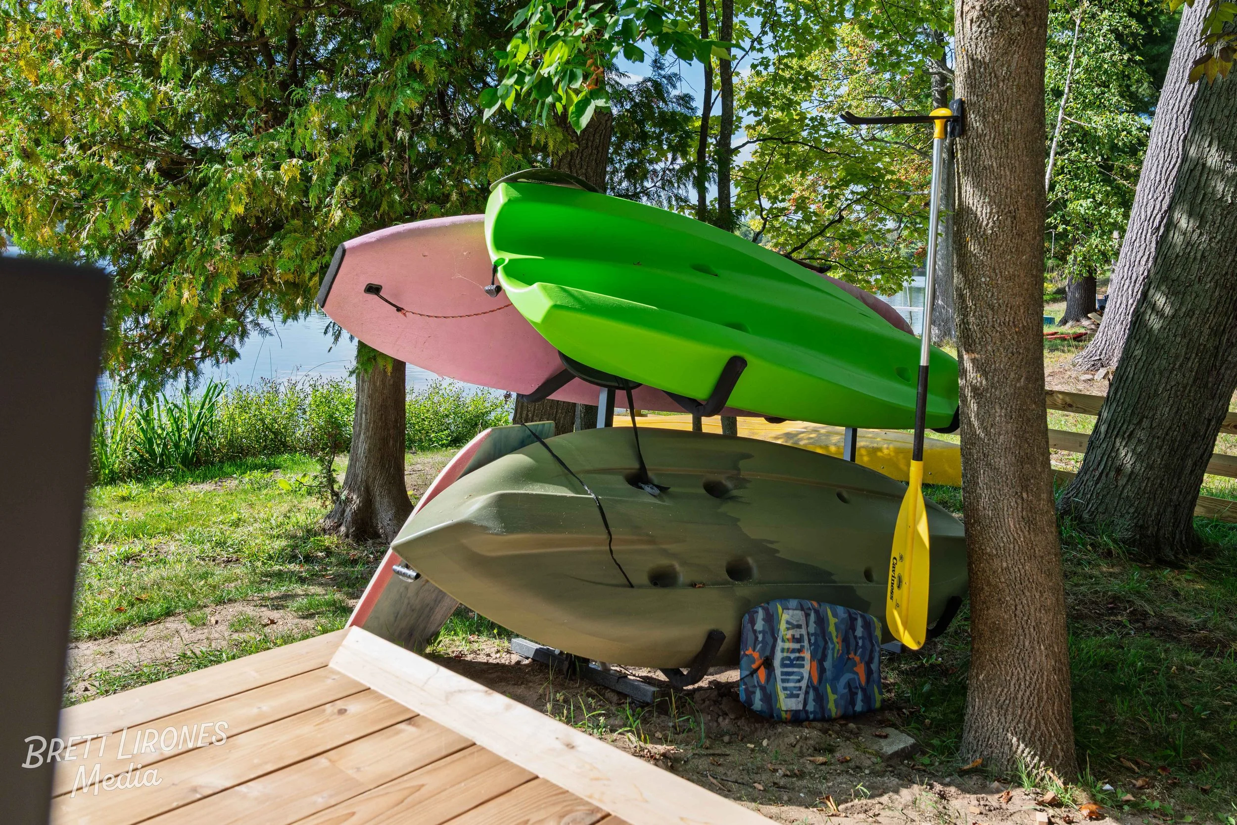A photographed outdoor scene showing three colorful kayaks, green, pink, and gray, stacked on a rack among trees near a body of water. There is a blue patterned life jacket or bag on the ground, and the area has a wooden deck in the foreground.