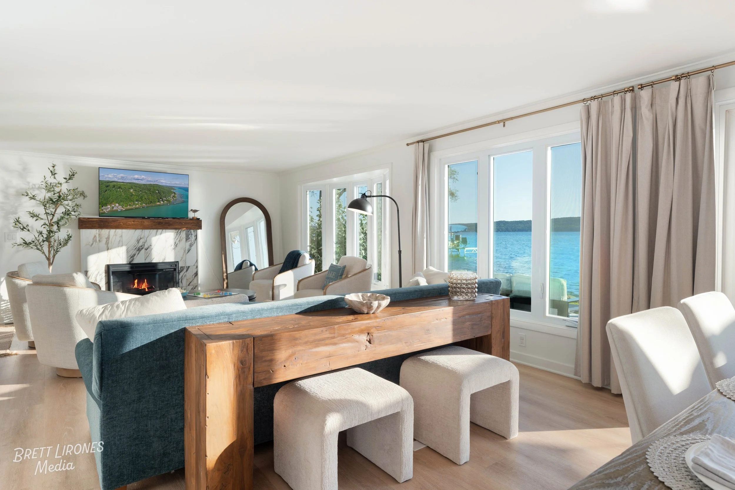 Bright living room with large windows overlooking a lake, featuring a fireplace, a flat-screen TV, seating area, wooden dining table, and neutral decor.