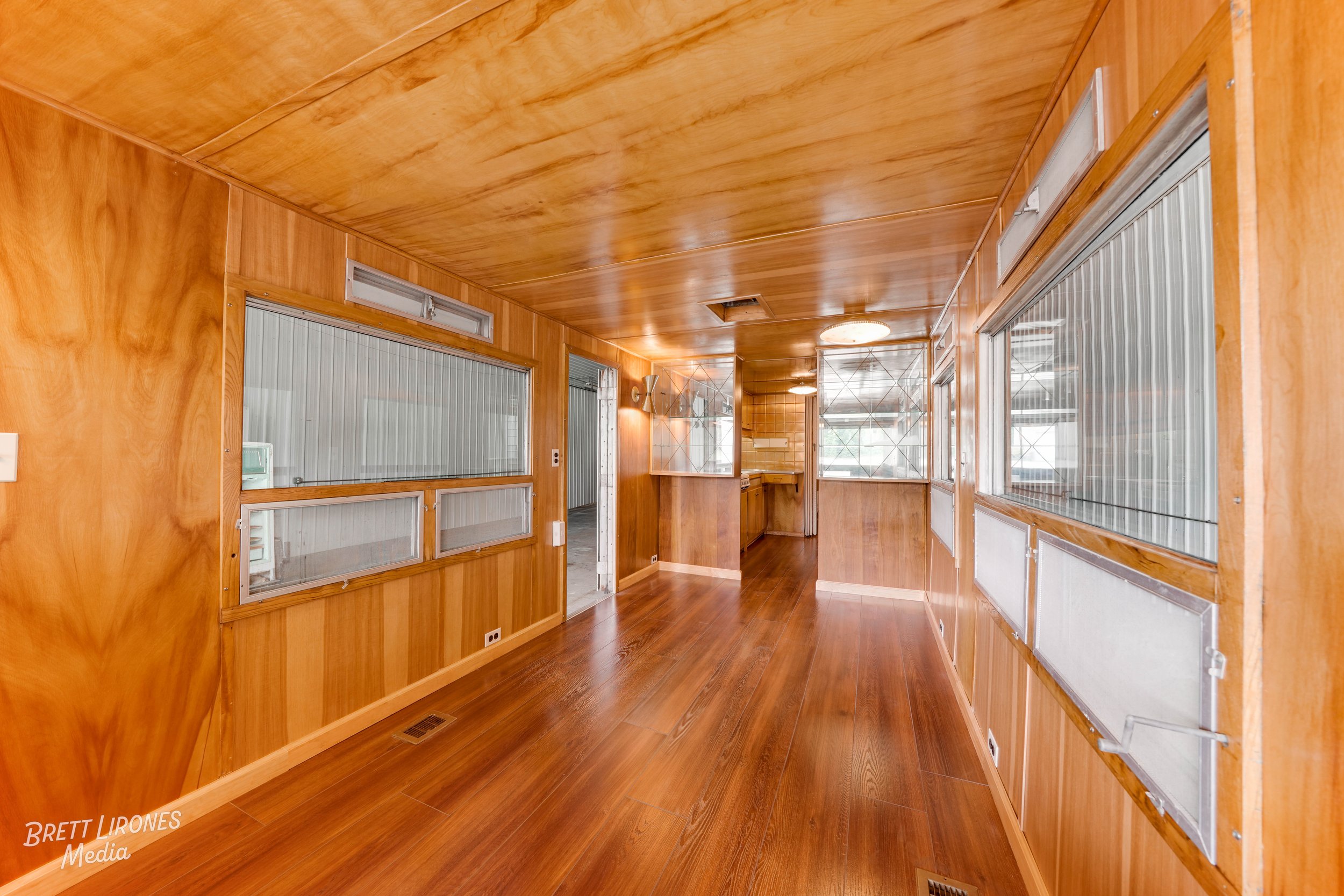 Wood-paneled interior room with large windows, hardwood flooring, a small kitchen area, and multiple ceiling lights.