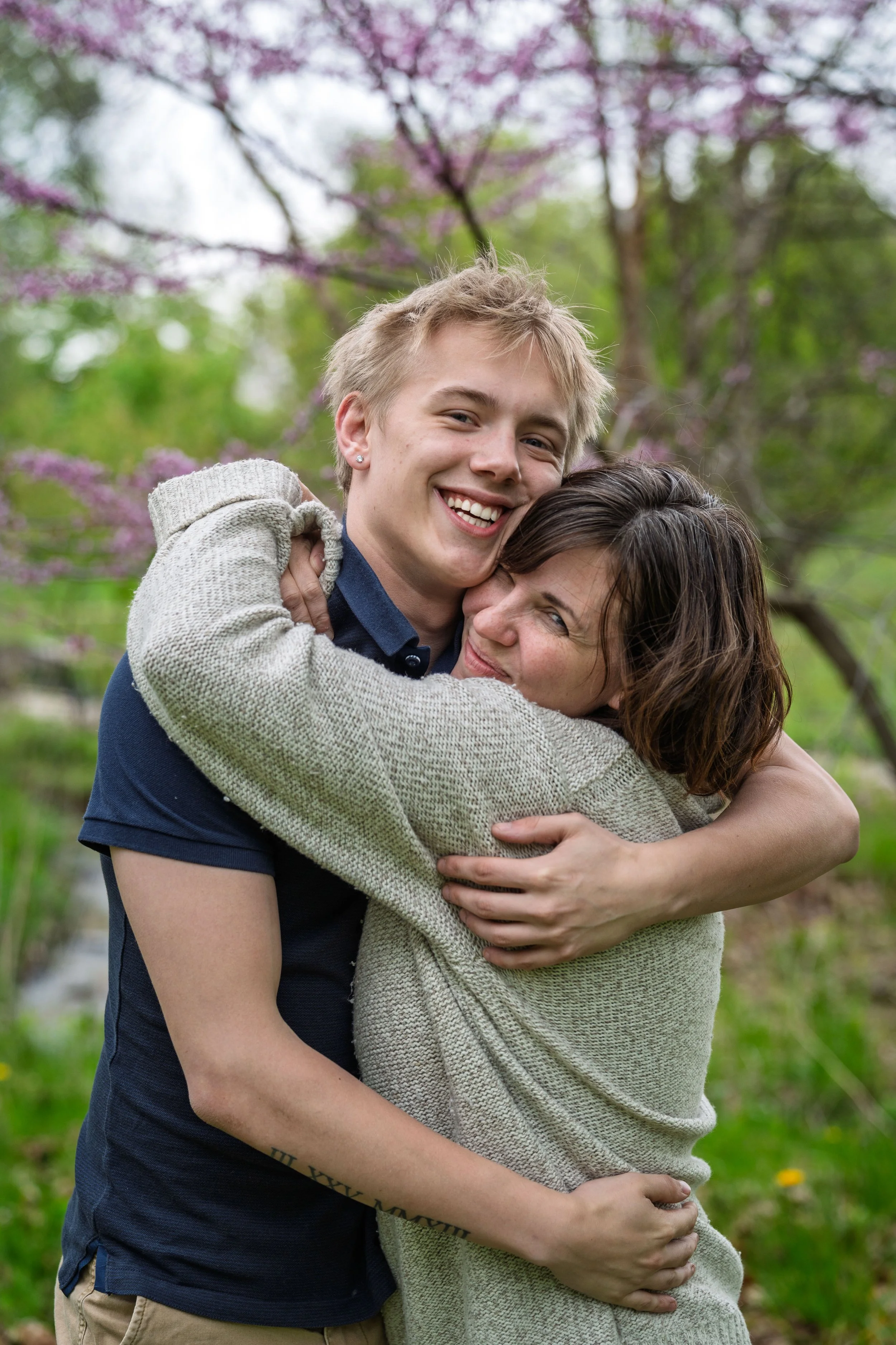 A young person and an older woman hugging outdoors with blooming pink trees in the background.