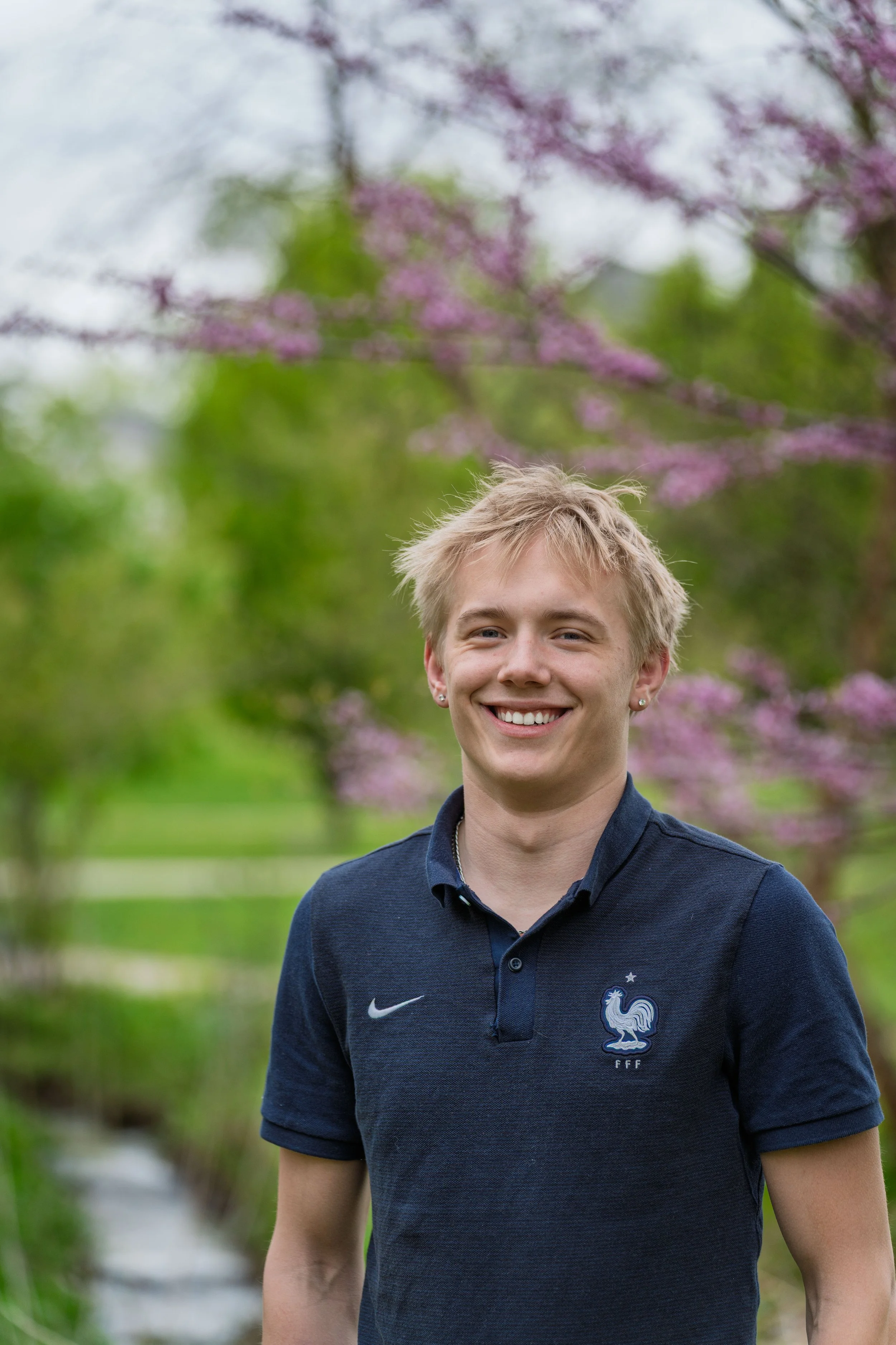 A young man with blonde hair smiling outdoors with pink flowering trees and green foliage in the background.