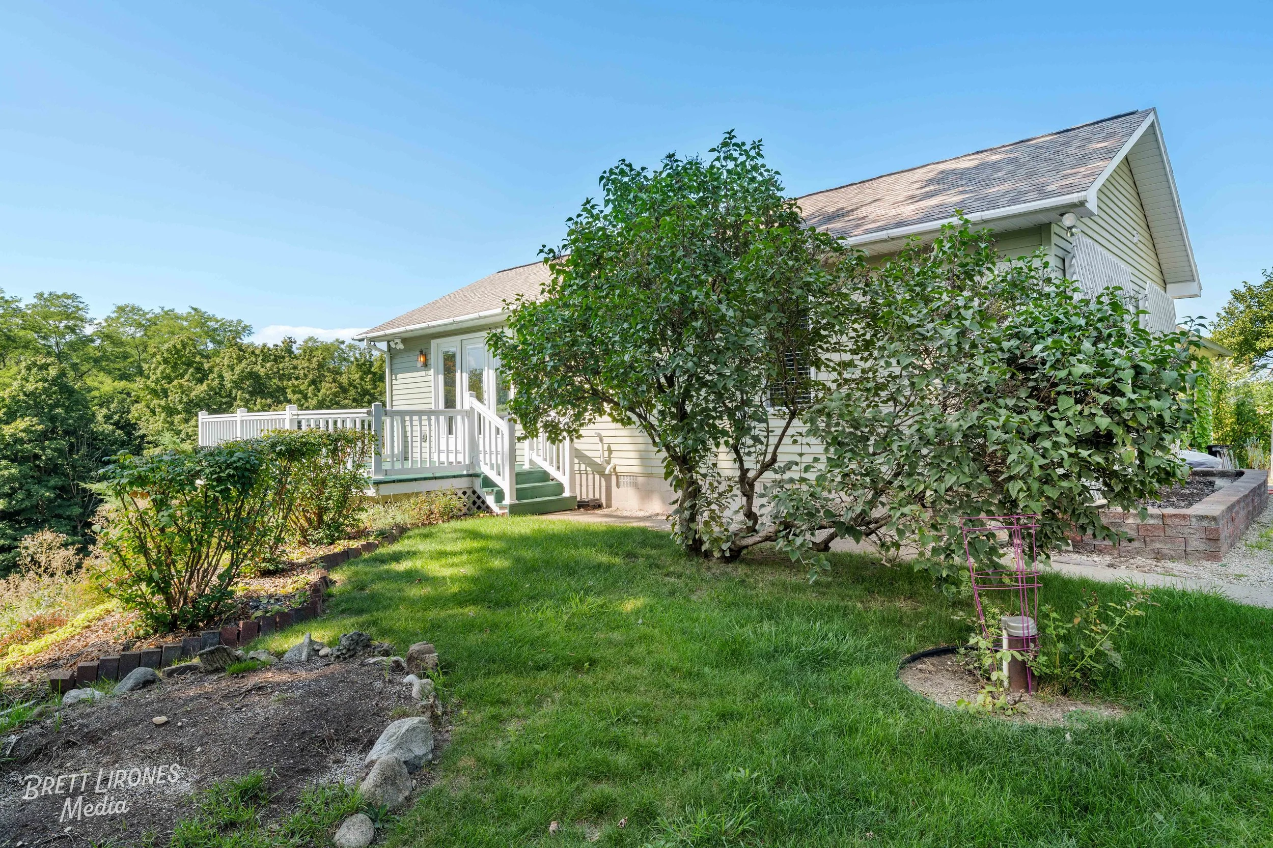Backyard view of a house with a small porch, surrounded by green bushes, trees, and a well-maintained lawn under a clear blue sky.
