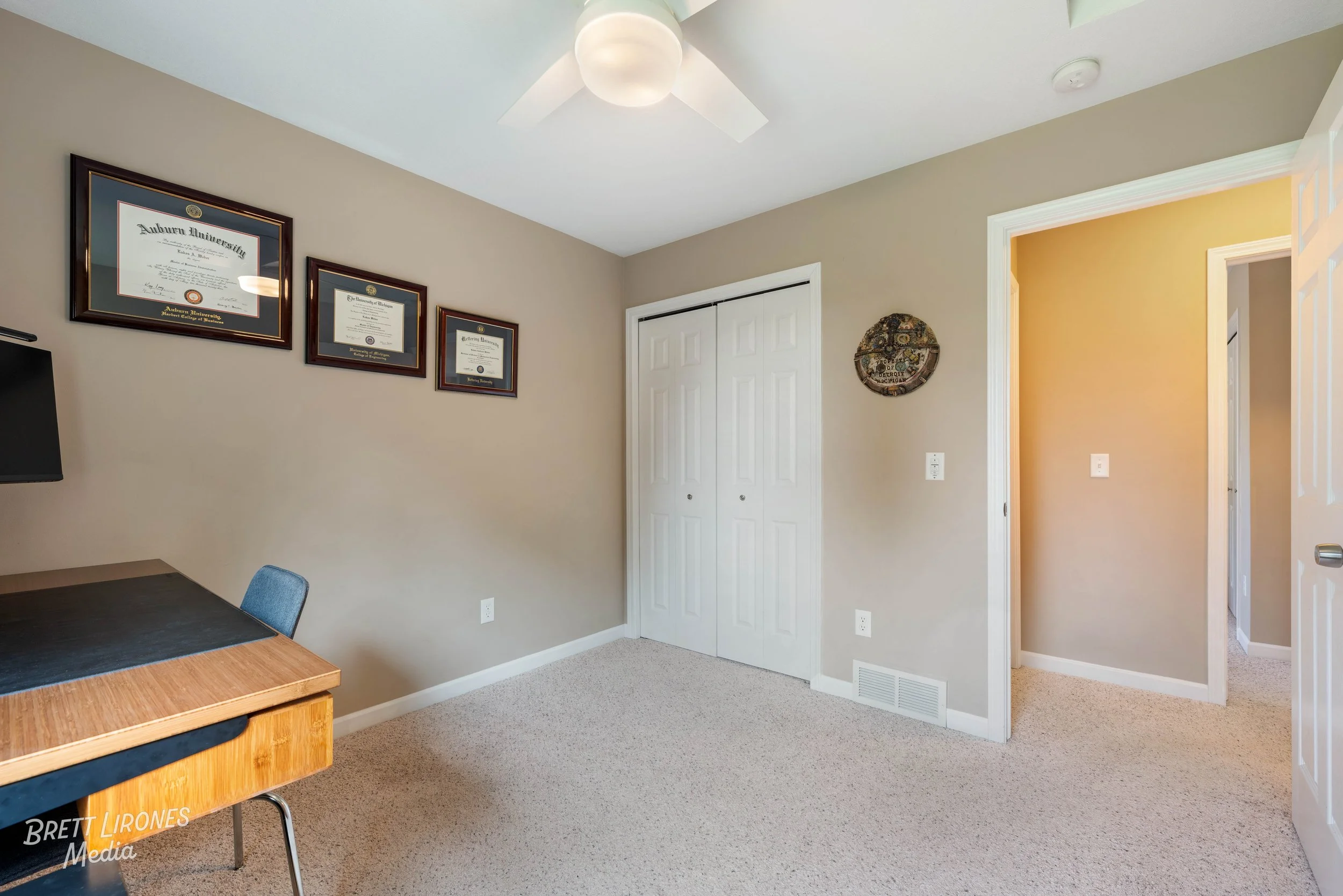 A room with beige walls, white closet doors, a ceiling fan with a light, and framed diplomas on the wall. There's a small desk with a chair and a circular wall decoration.