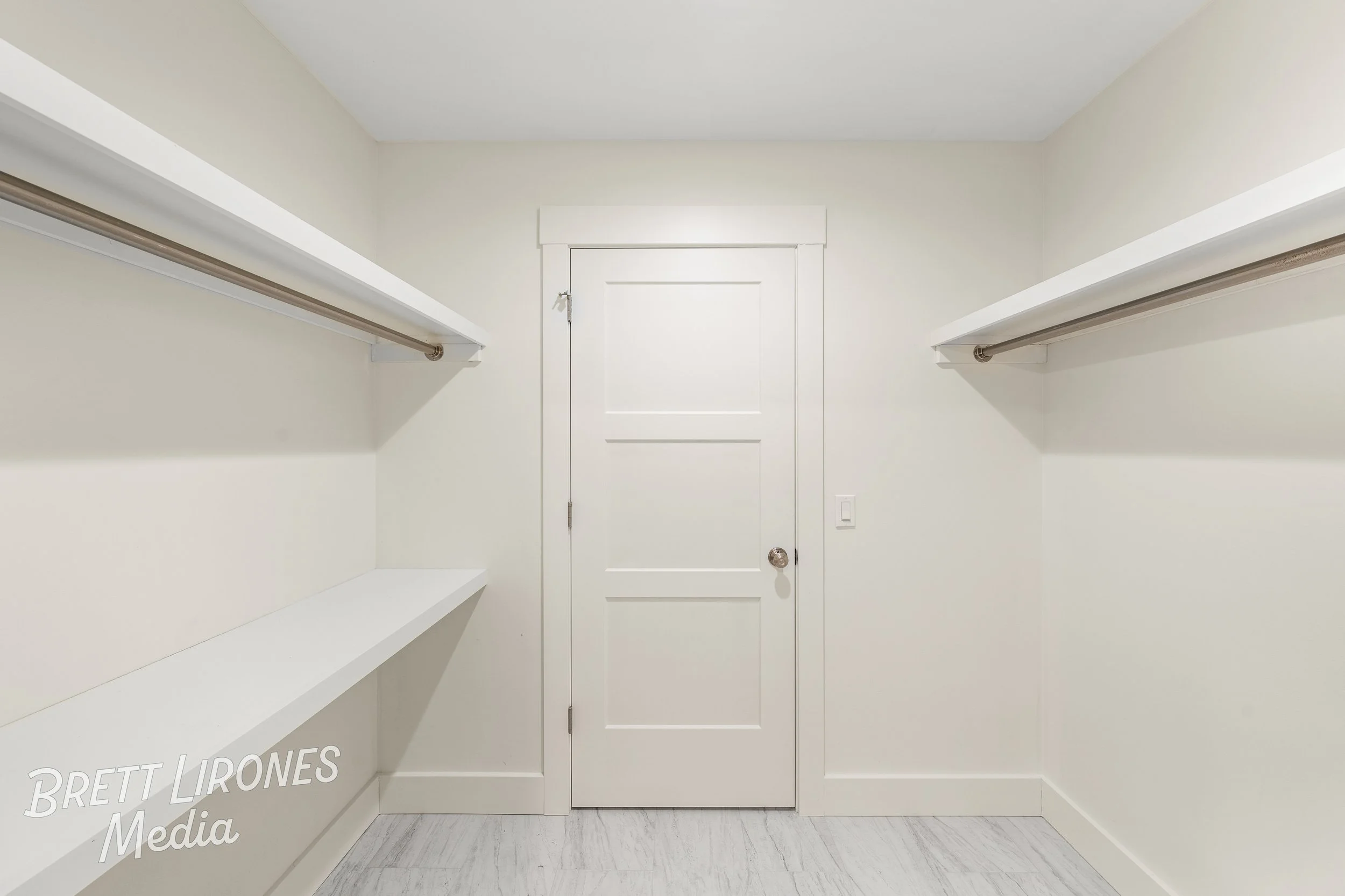 Empty walk-in closet with white shelves on both sides, white door in the center, and light-colored flooring.