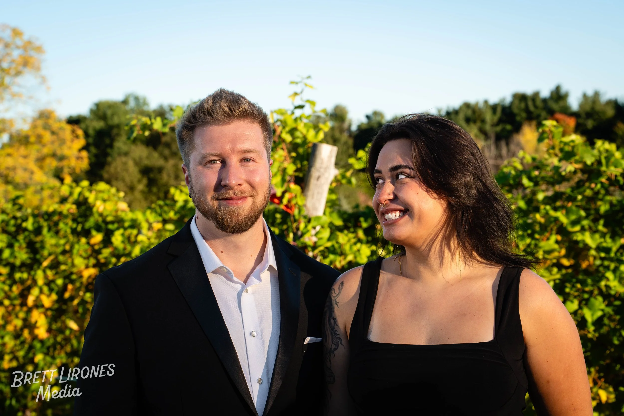 A smiling man in a black suit and a woman with dark hair and a nose ring, smiling at each other outdoors with green bushes and trees in the background.