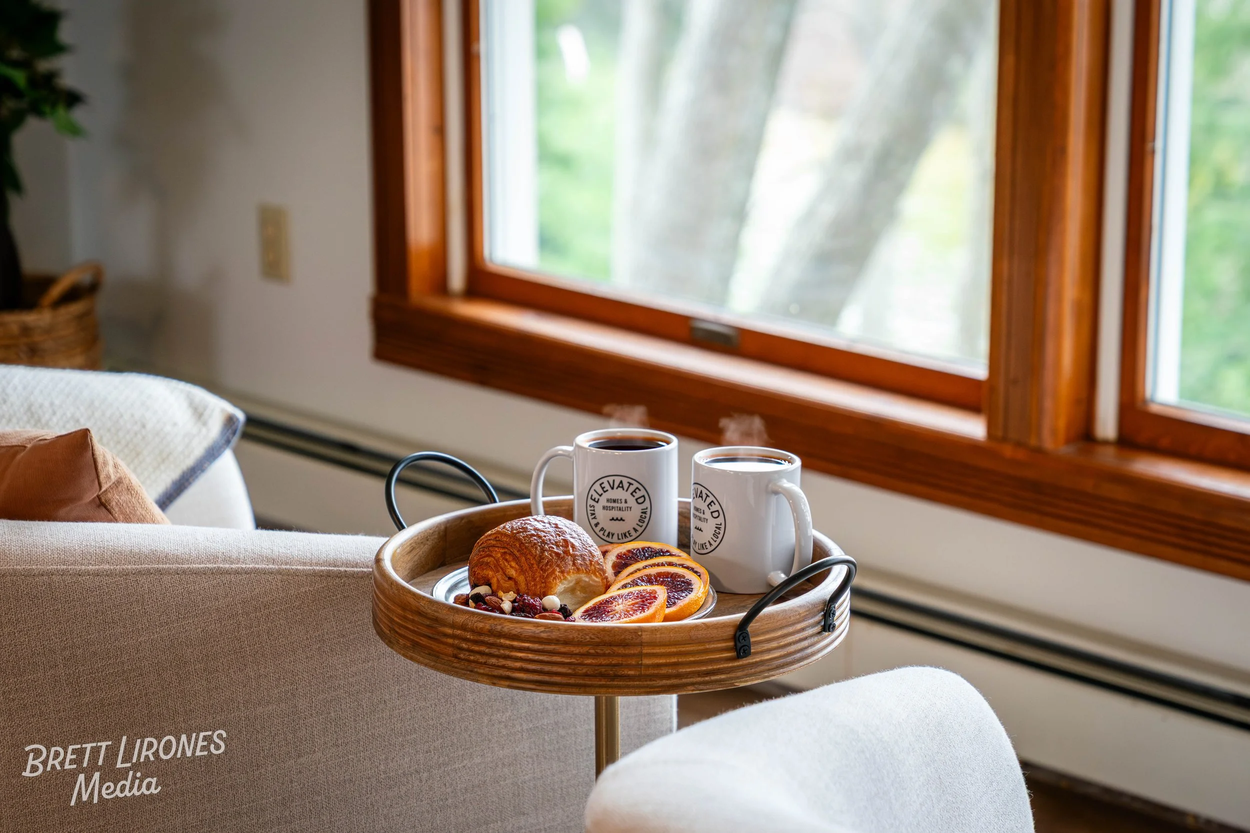 Two steaming mugs with a logo, a croissant, sliced blood oranges, and assorted nuts on a wooden tray in front of a window with a view of green trees.