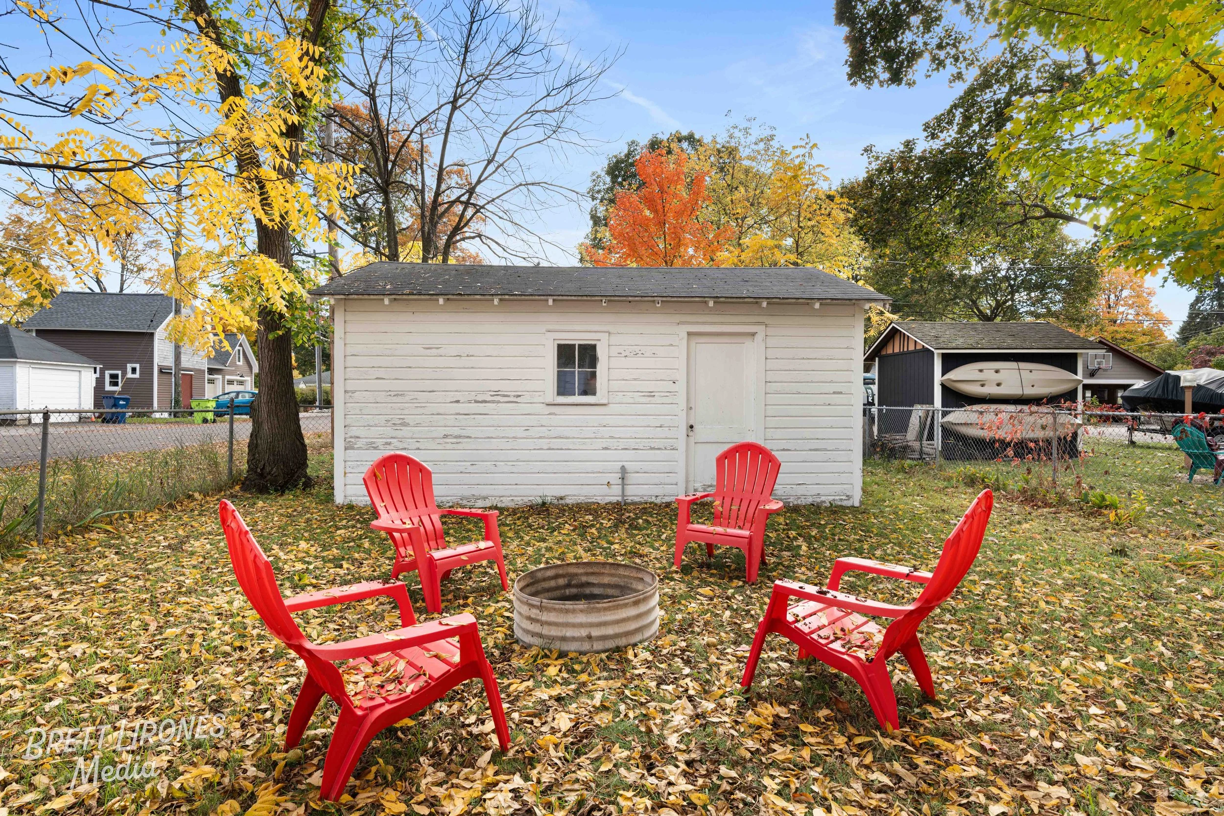 Backyard with four red chairs around a fire pit and fallen autumn leaves, white shed in background, autumn trees with yellow, orange, and green leaves, blue sky.