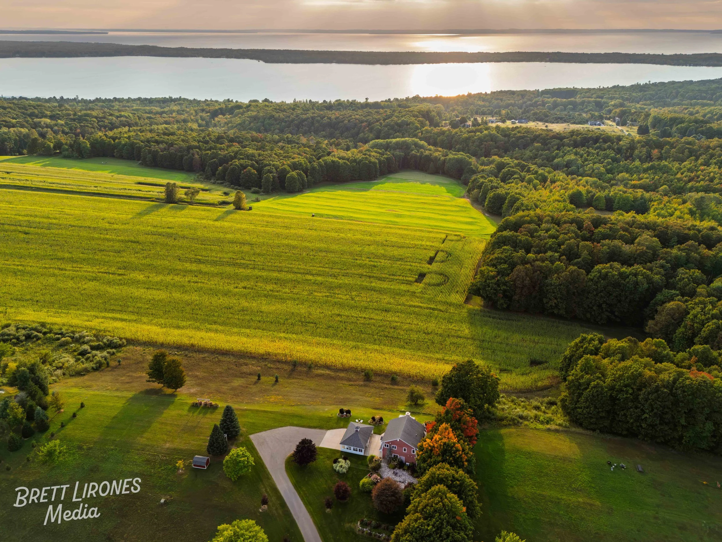Aerial view of a farm with a house and barn surrounded by green fields and trees, near a large body of water at sunset.