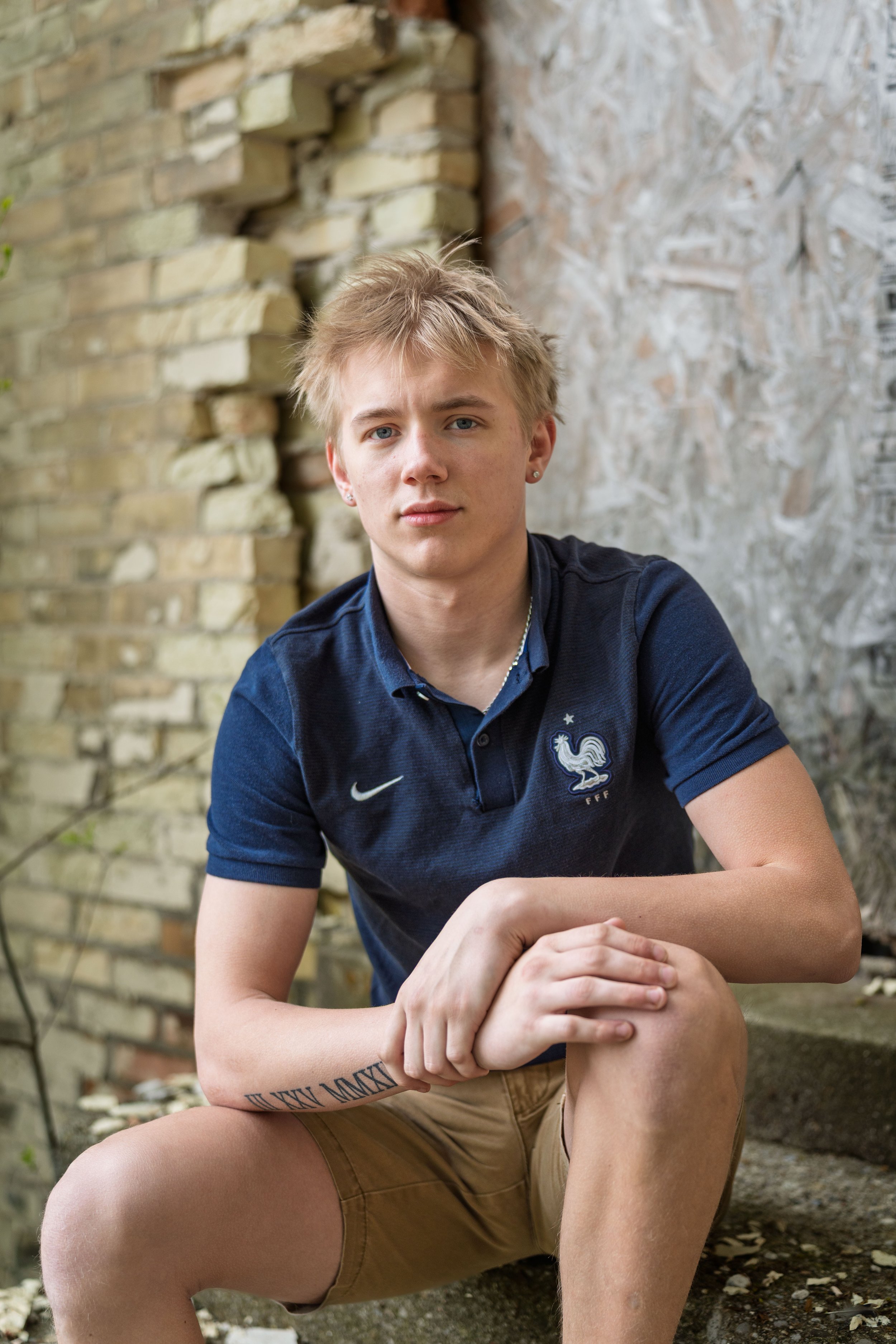 A young man with blonde hair and light skin sits on outdoor steps near a brick wall. He wears a navy blue polo shirt with a French soccer emblem and tan shorts, and looks into the camera with a neutral expression.
