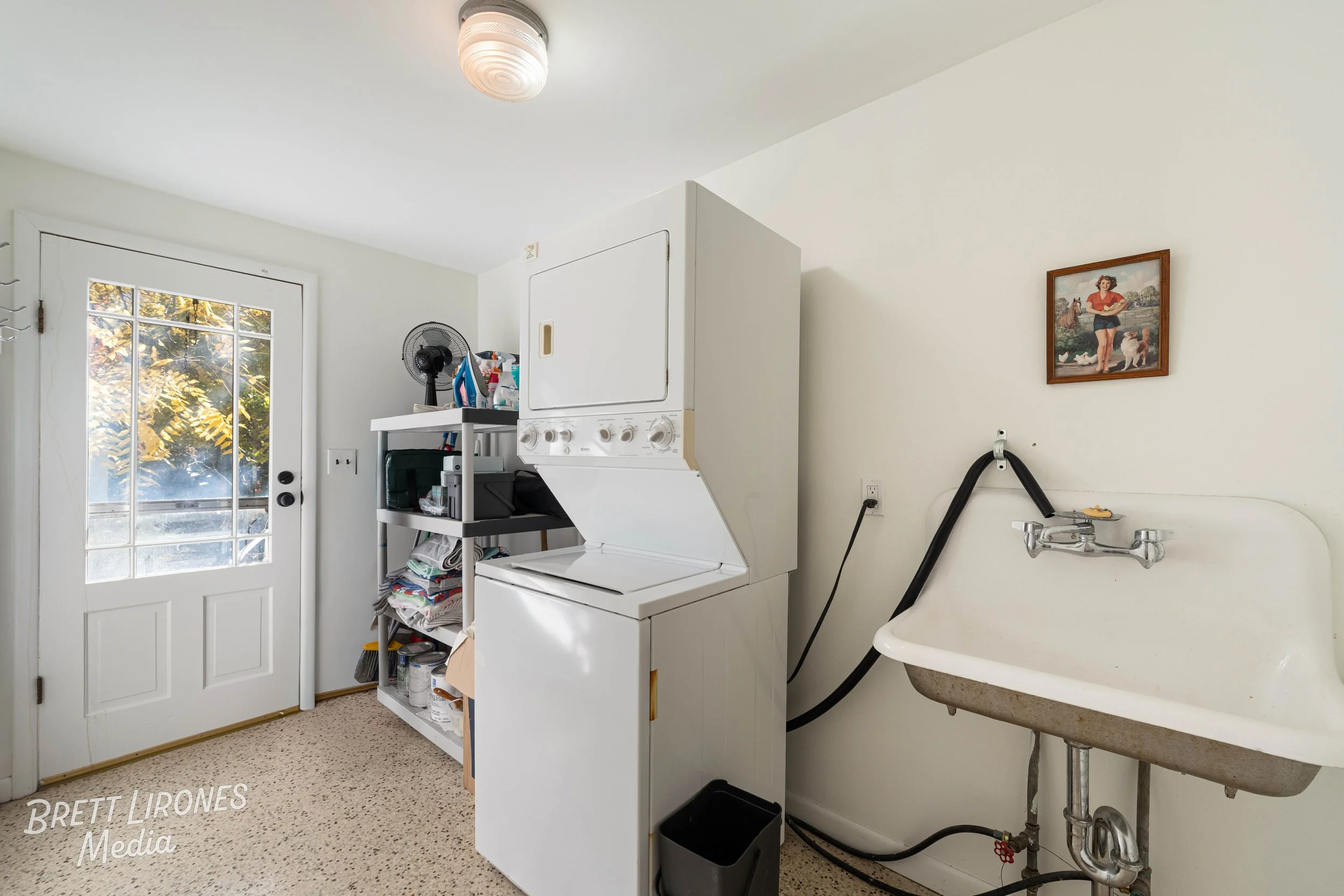 Laundry room with a stacked washer and dryer, a shelf with miscellaneous items, a small fan, a door with a window, and a utility sink with exposed pipes and a framed picture on the wall.