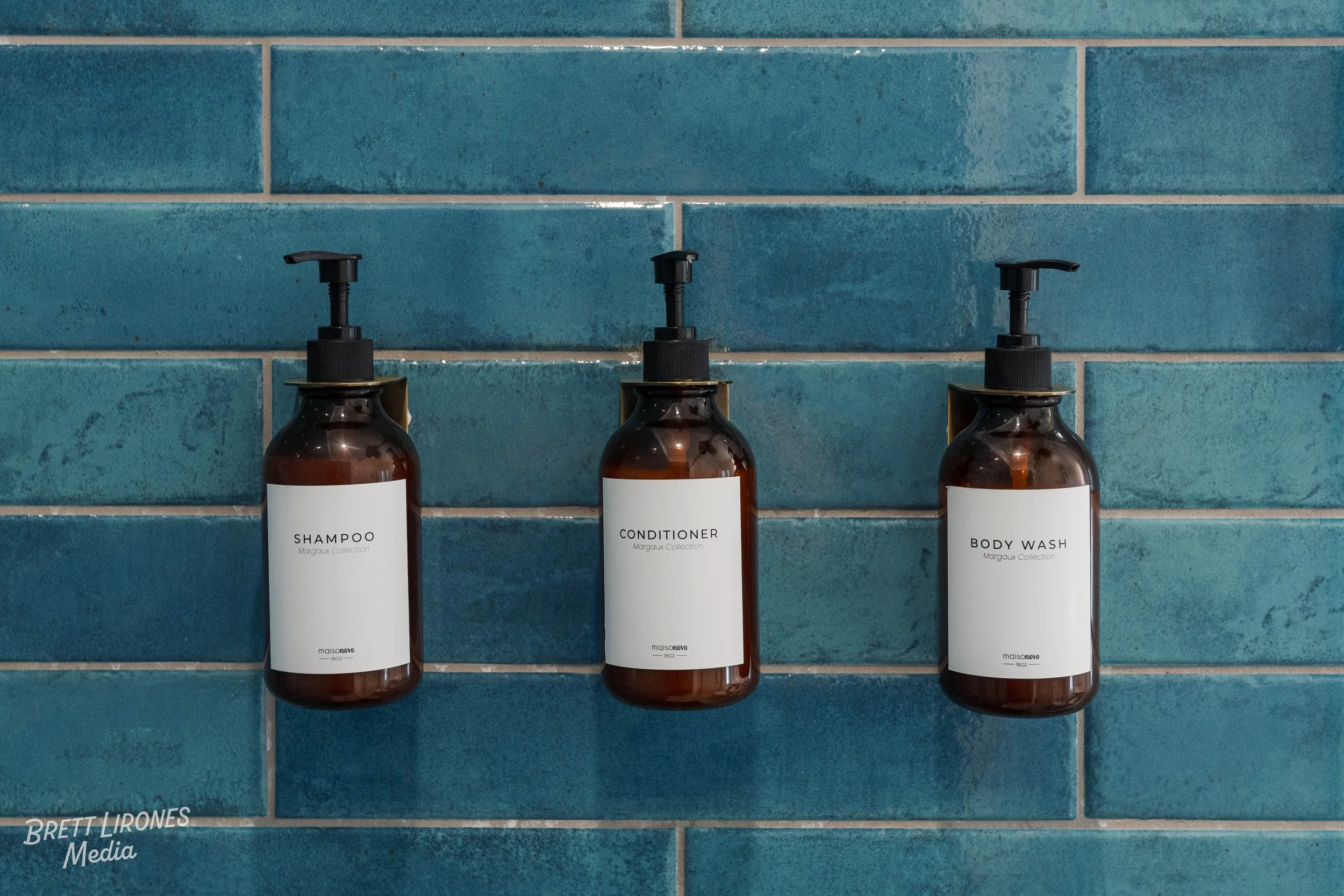 Three brown bottles with black pump dispensers labeled shampoo, conditioner, and body wash, mounted on a blue tiled wall.