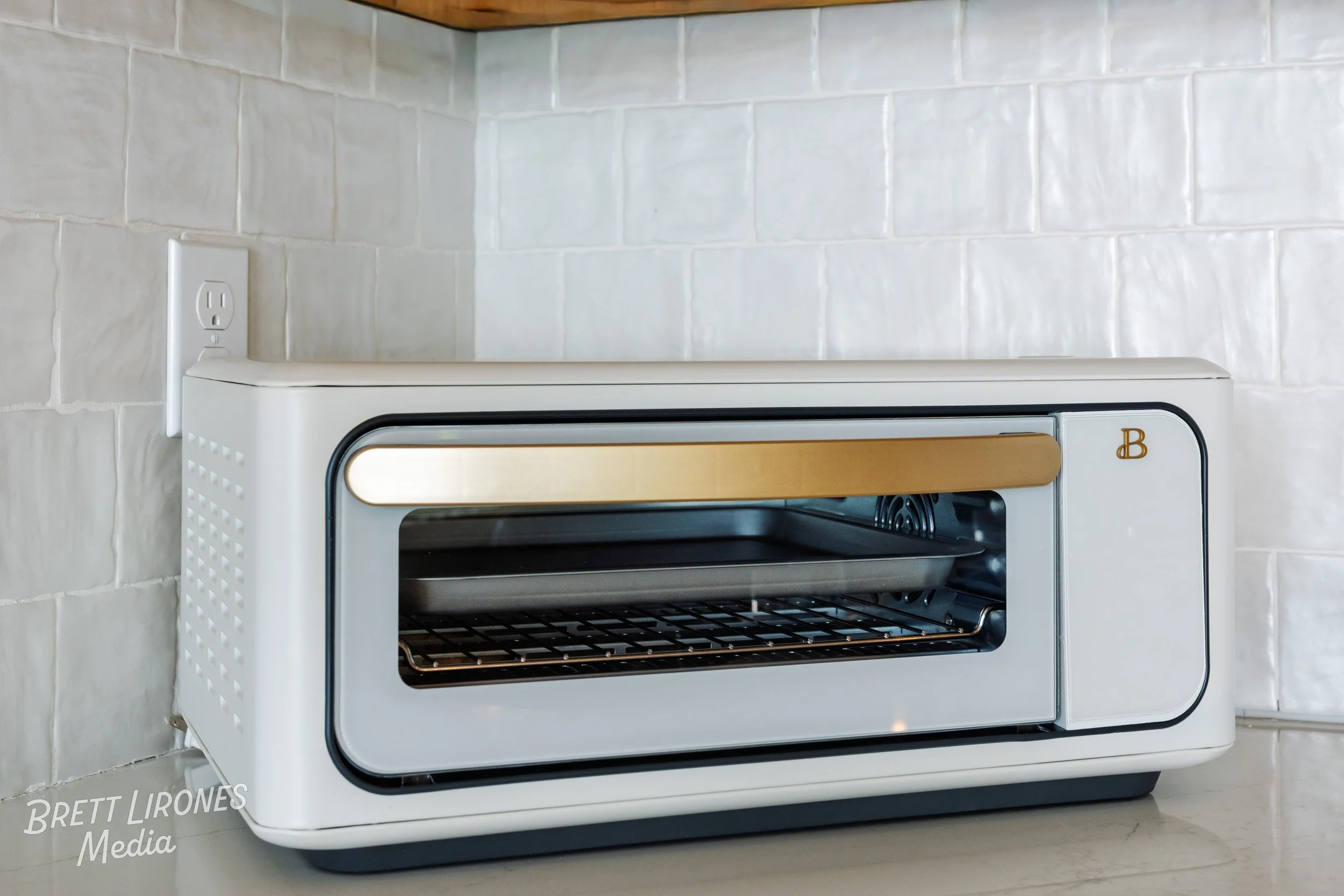 A small, white toaster oven with a gold handle and a glass door, placed on a kitchen countertop near an electrical outlet.