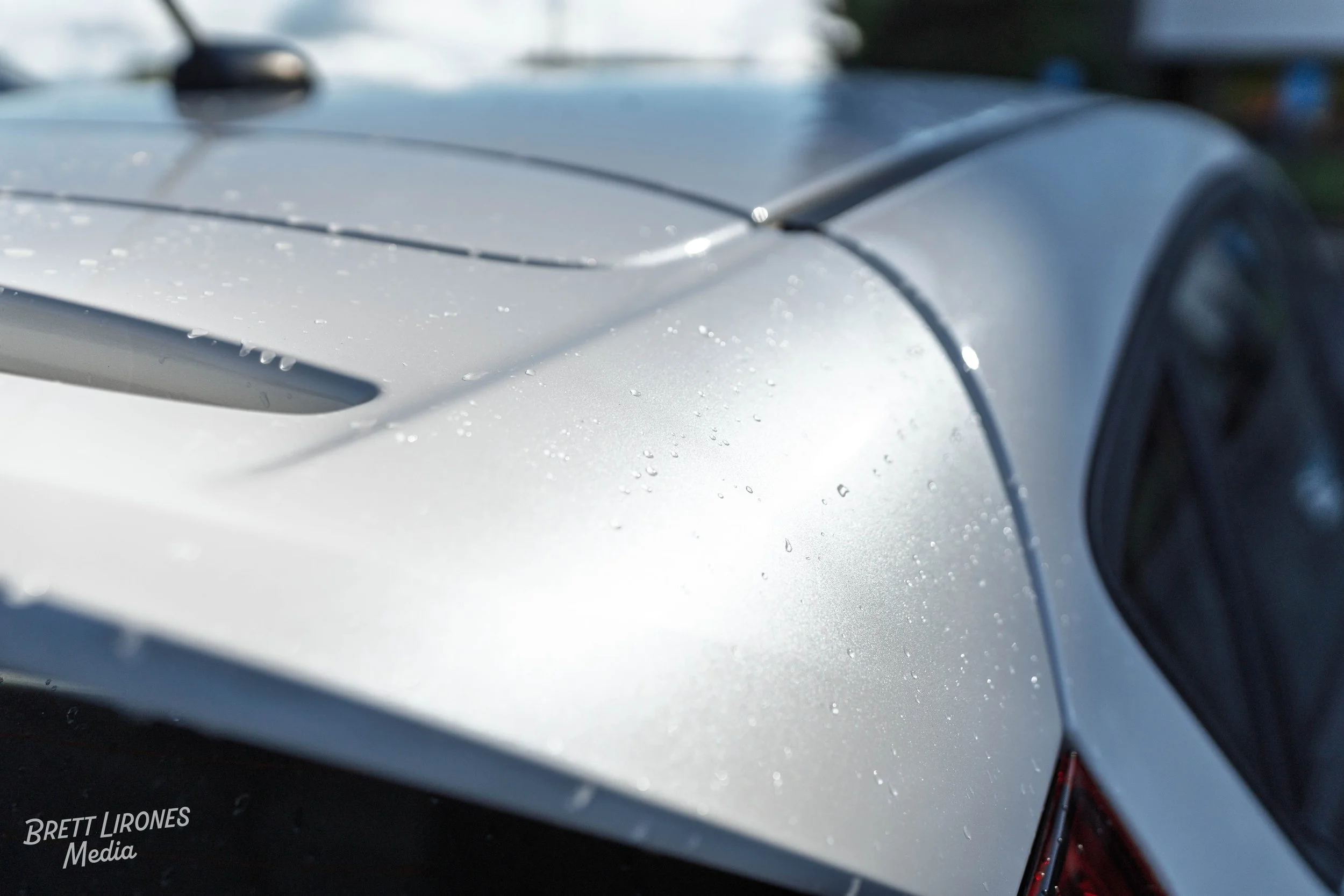 Close-up of a silver car's rear with water droplets on the surface, showing part of the rear window and roof.
