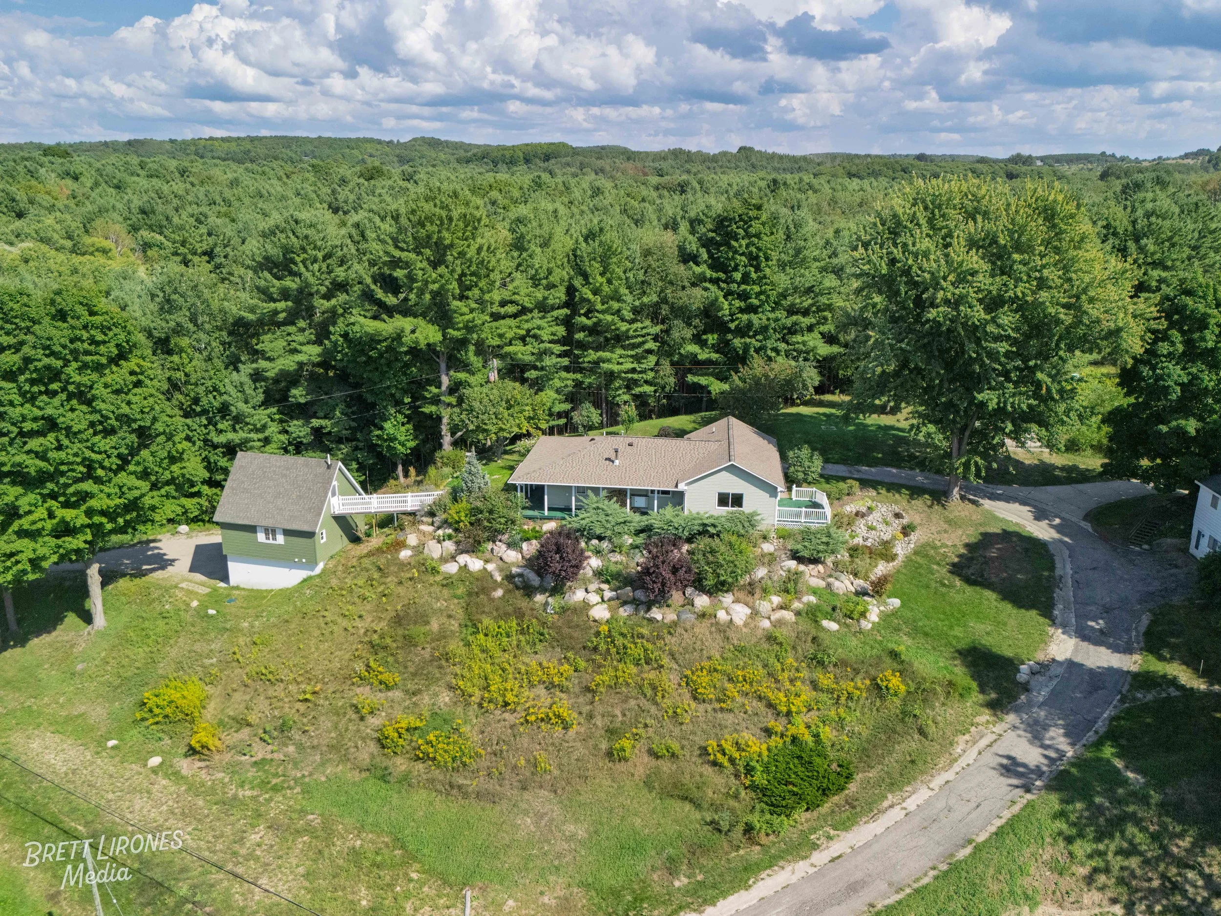 Aerial view of a rural property with a main house, a small separate building, a winding driveway, and lush green trees in the background, under a partly cloudy sky.
