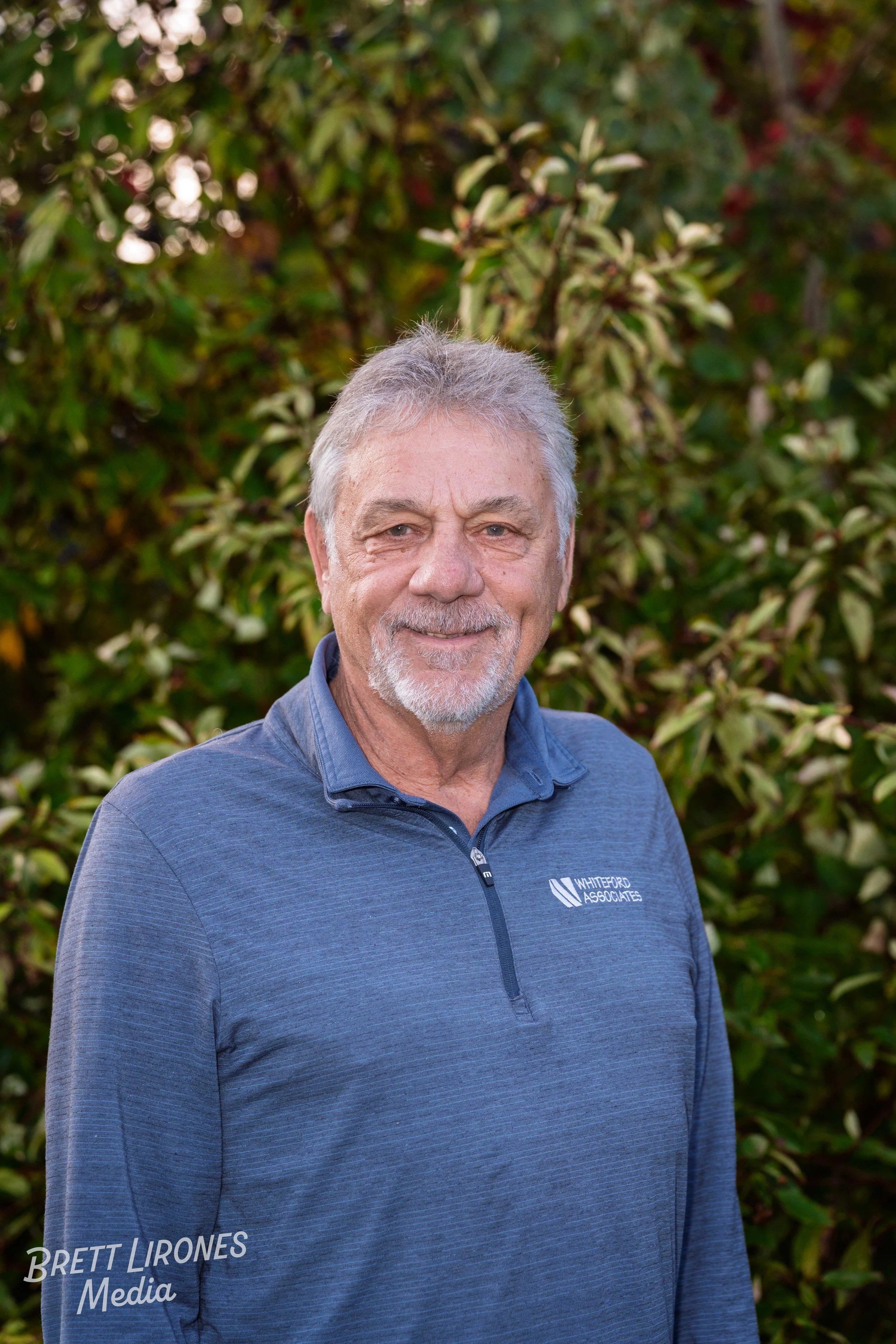 A smiling older man with gray hair and a beard, wearing a blue quarter-zip sweatshirt embossed with 'Whiteford Associates,' standing outdoors in front of green and red foliage.