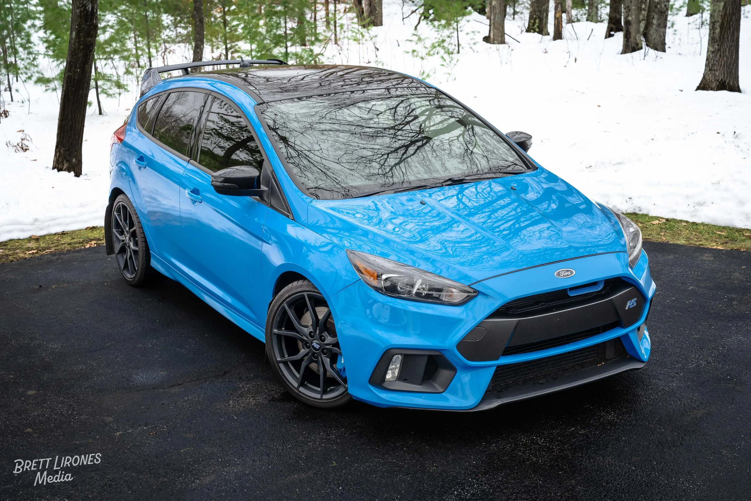 Blue Ford Focus RS parked on a dark asphalt surface with snow and trees in the background.