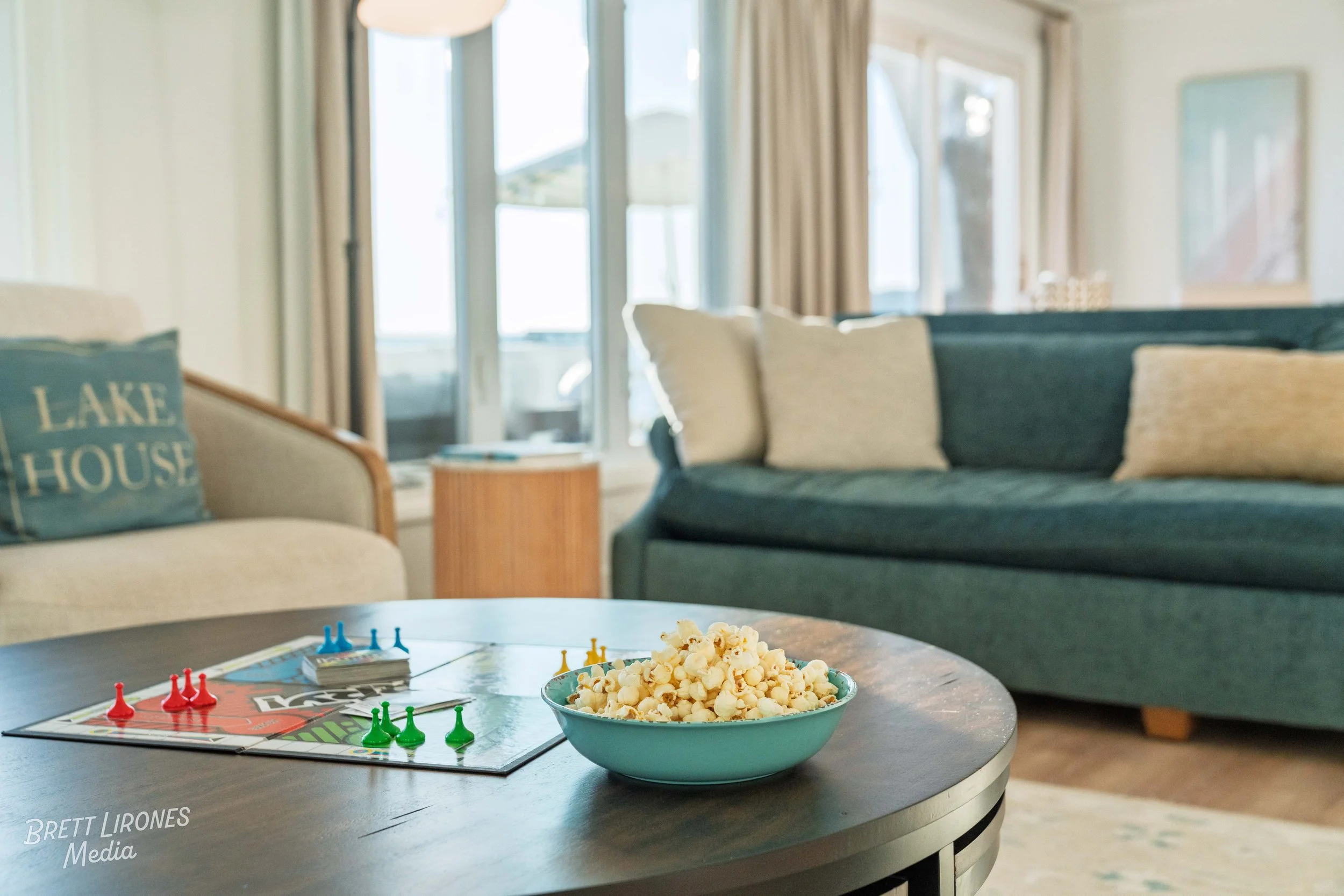 A close-up of a round wooden coffee table with a bowl of popcorn and a Monopoly game with colored player pieces on it, in a bright living room with beige and blue sofas and large windows with curtains.