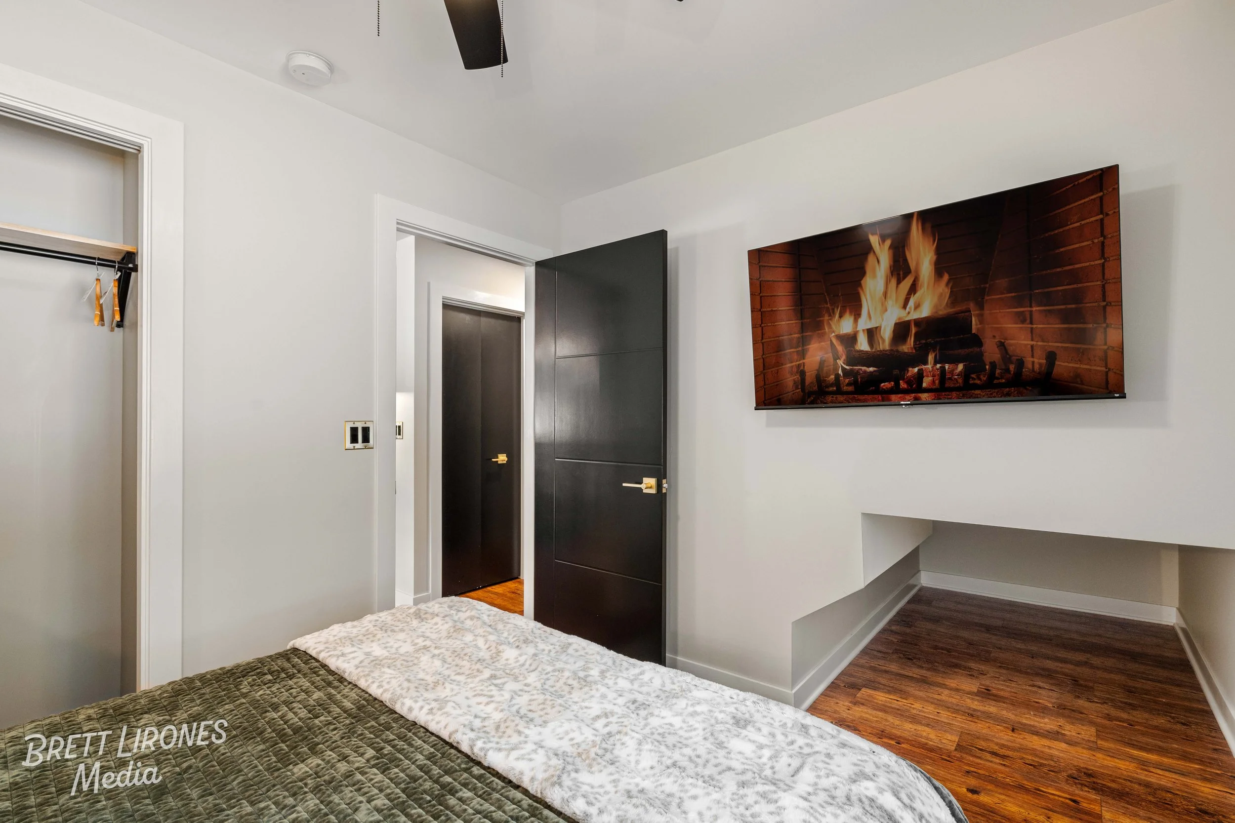 Bedroom with a bed, wall-mounted TV showing a burning fire, dark door, open closet, white walls, hardwood floor.
