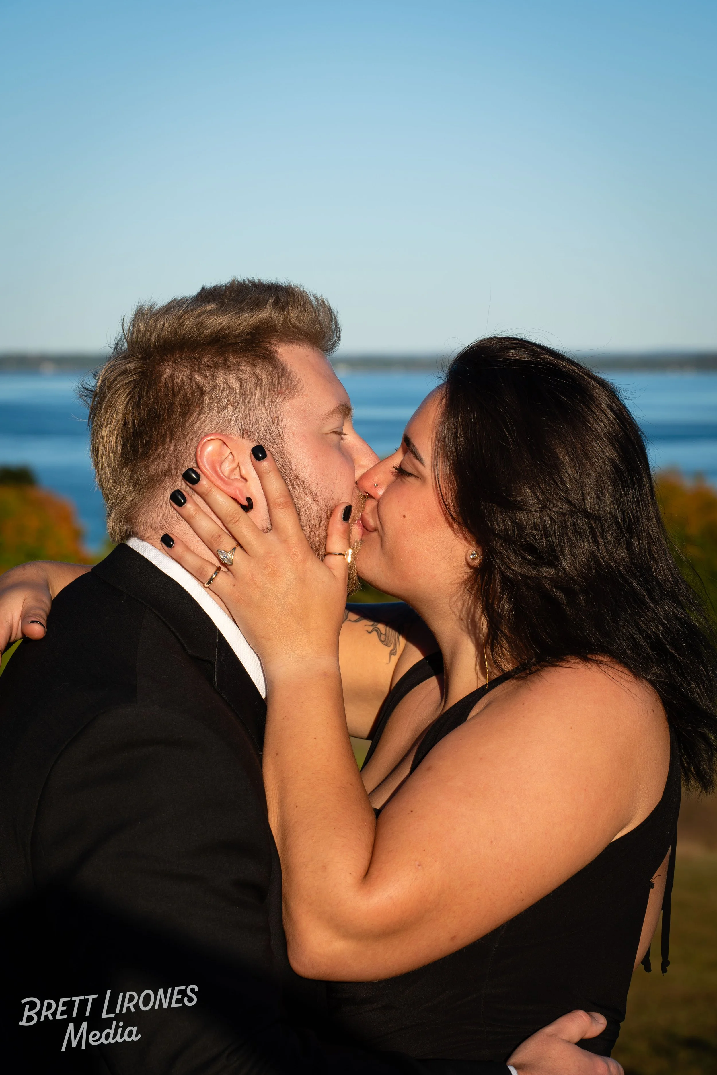 A couple sharing a kiss outdoors near a body of water under a clear blue sky.