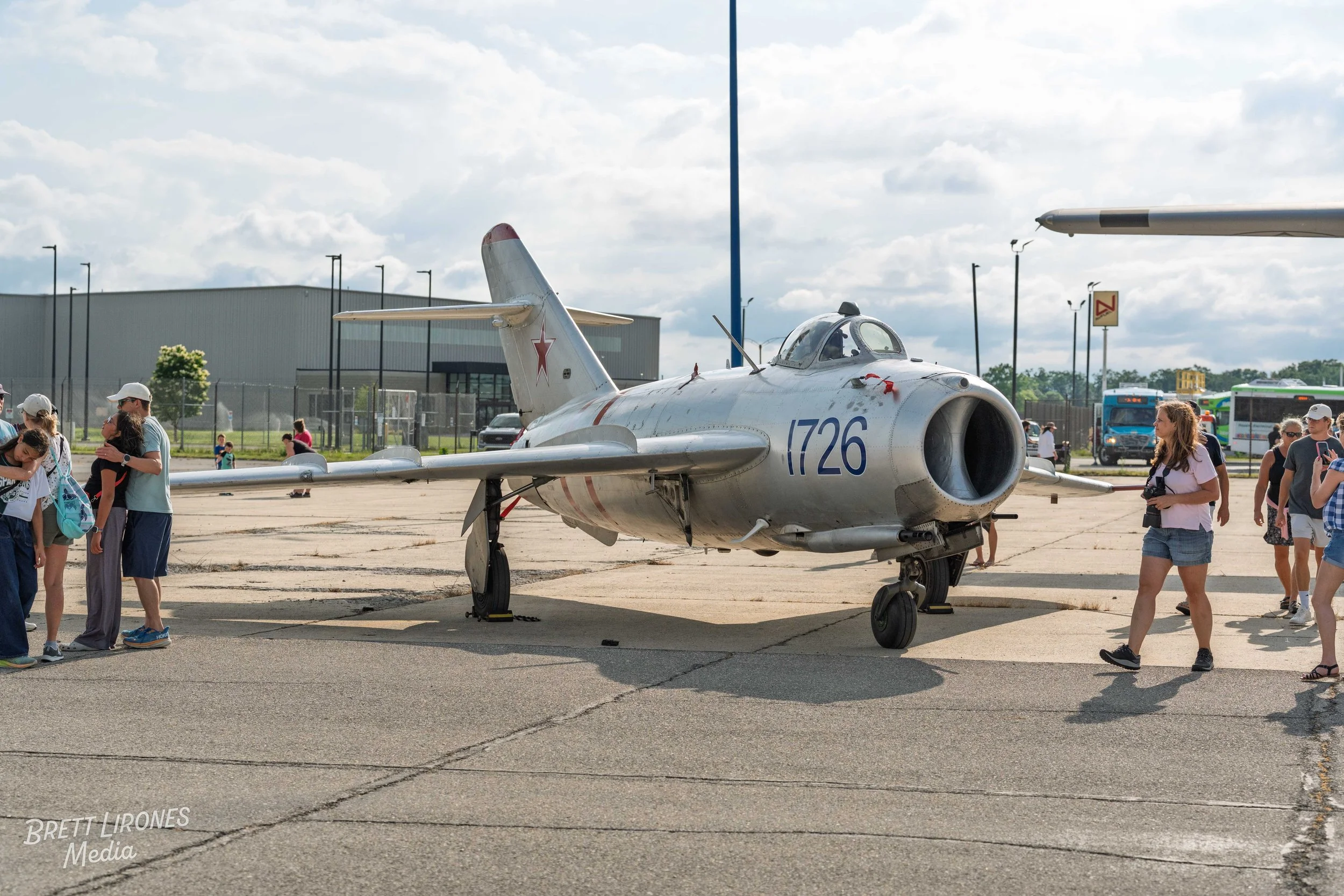 A vintage military jet aircraft on display outdoors with children and adults viewing it. The aircraft is silver with number 1726 and Red Star insignia on the tail, parked on a concrete surface under a partly cloudy sky.