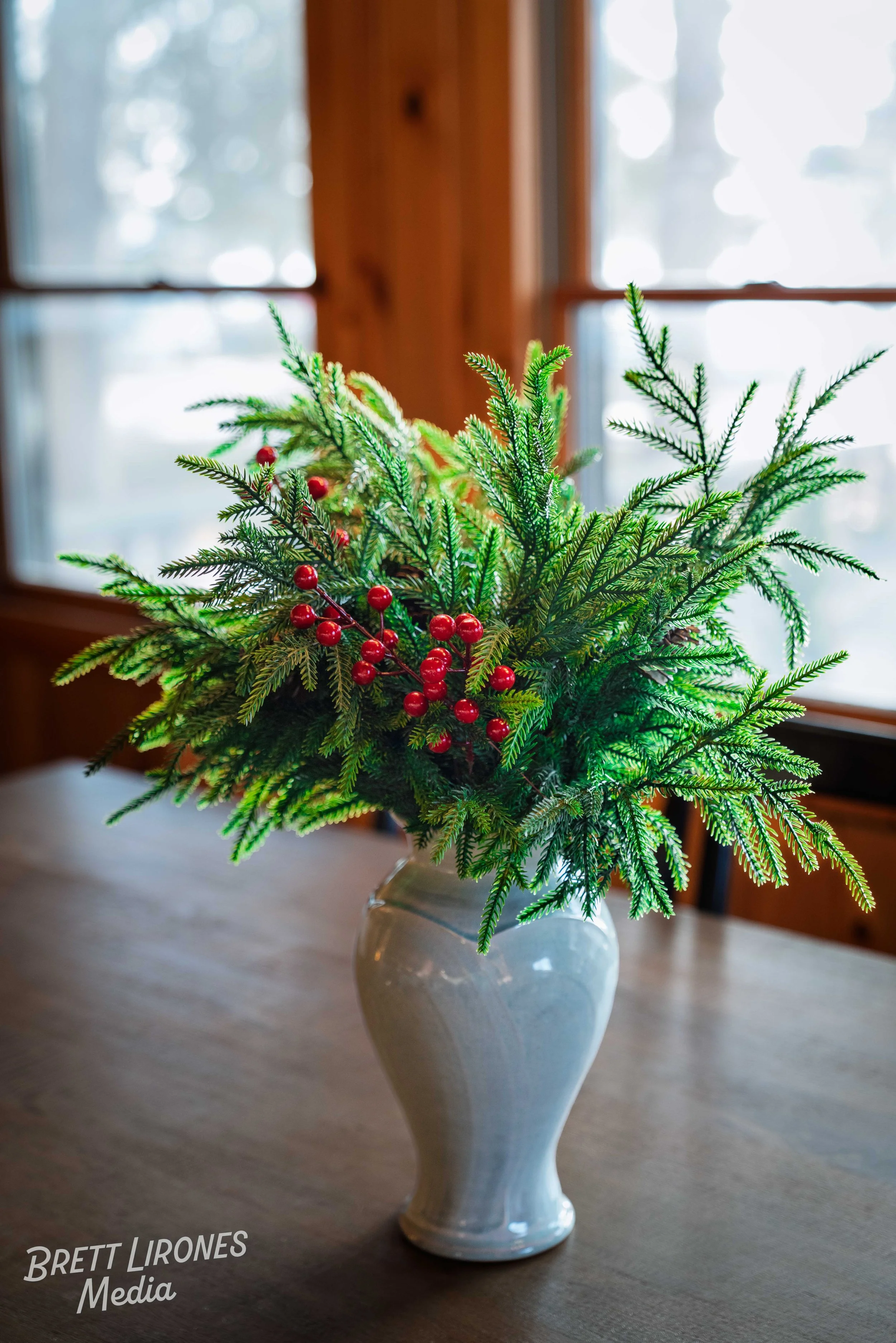A white vase filled with green pine branches and red berries, placed on a wooden table near a window.