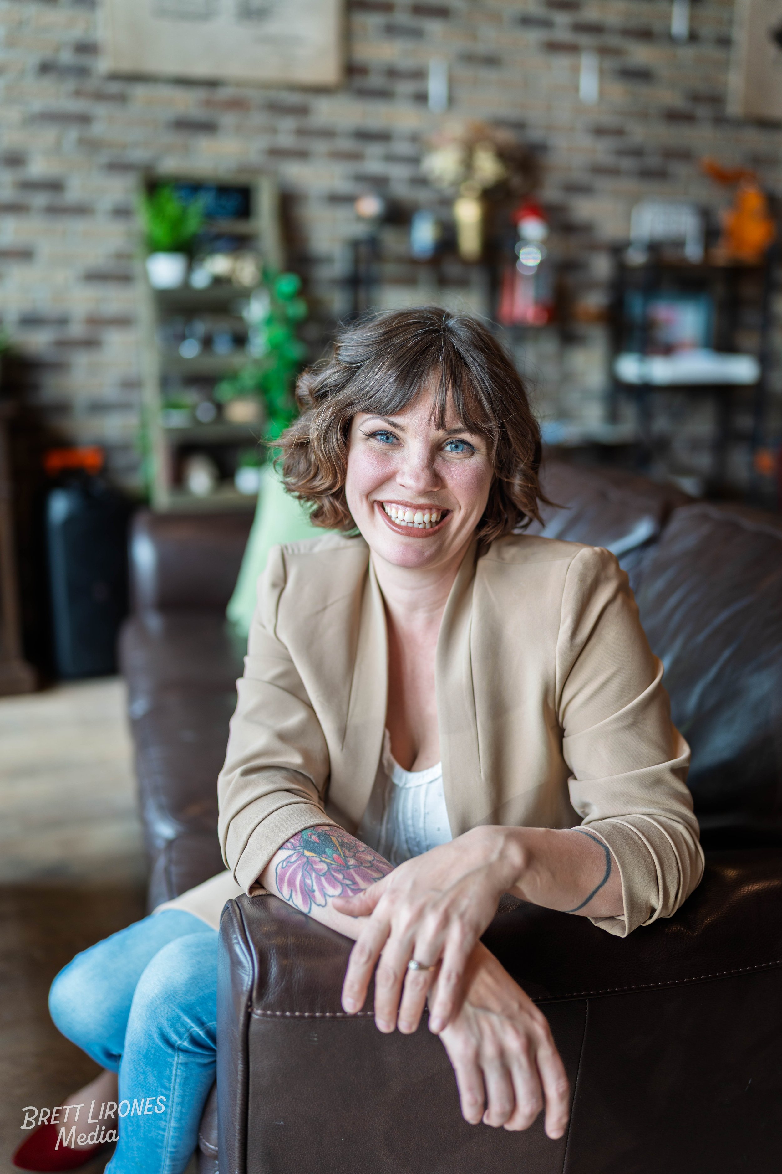 A smiling woman with curly brown hair and tattoos on her arms sitting on a leather couch in a cozy, decorated room.