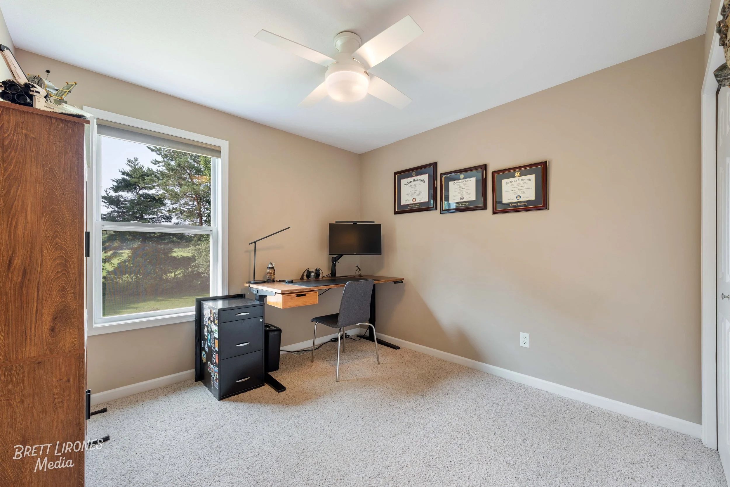 Home office with a window, desk, chair, computer monitor, framed certificates on the wall, ceiling fan, and a wooden cabinet.