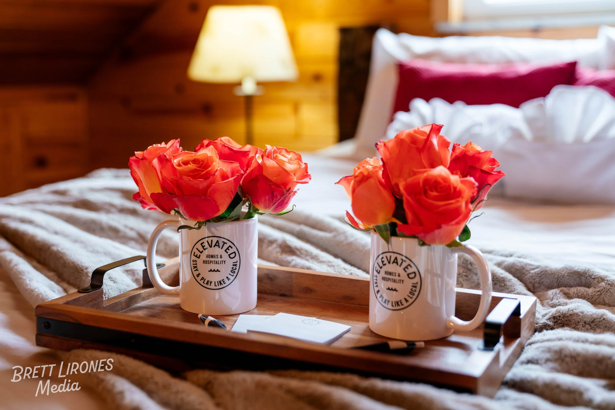 Two white mugs with black text and logo, each filled with pink and orange roses, placed on a wooden tray on a bed with a beige blanket, in front of bed pillows and a lamp in a wooden room.