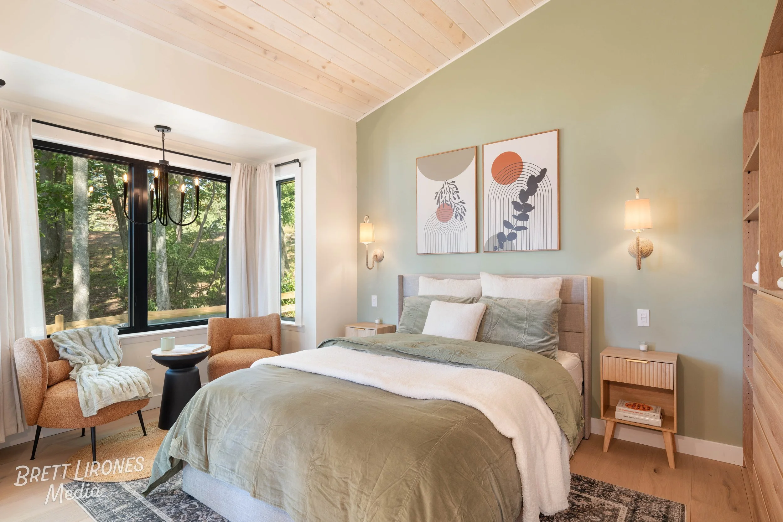 A bedroom with a large window overlooking trees, a bed with green and white bedding, two art pieces on the green wall, two small wooden nightstands, and two wall lamps.