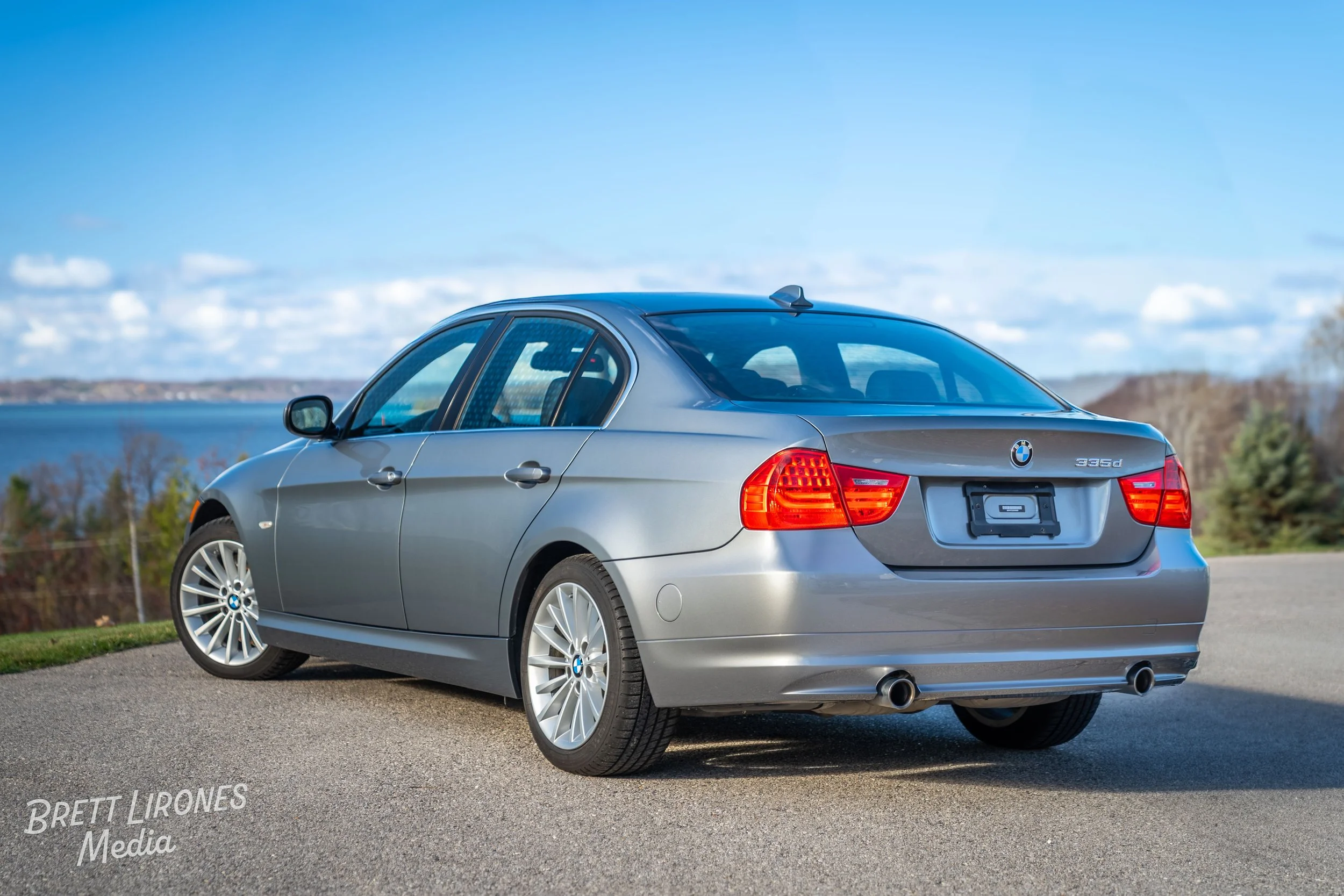 Silver BMW 3 Series sedan parked outdoors on a paved surface with a scenic background of water, trees, and a partly cloudy sky.