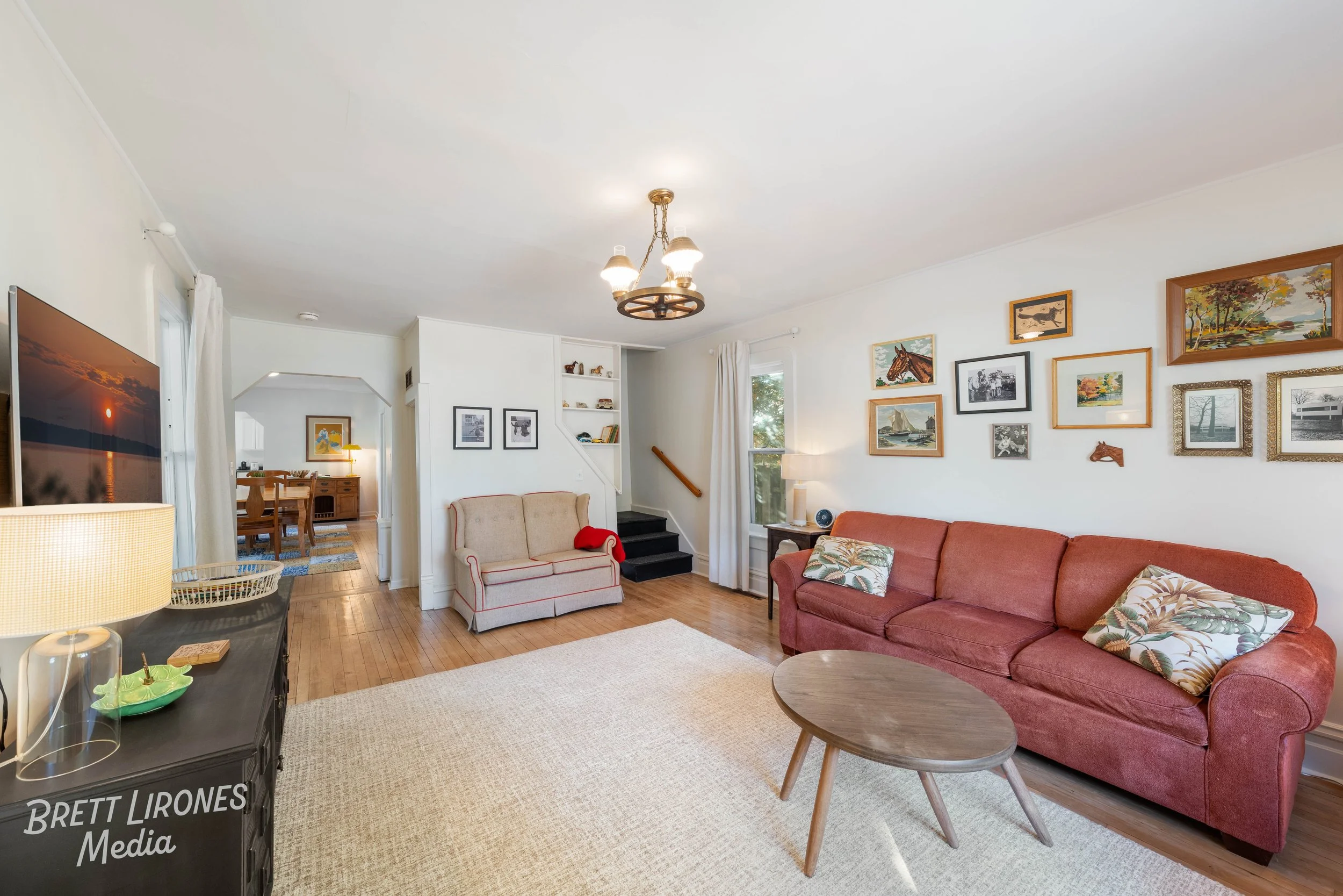 Living room with a red sofa, beige armchair, wooden coffee table, and framed artwork on white walls. There is a chandelier, a side table with a lamp, and a television.