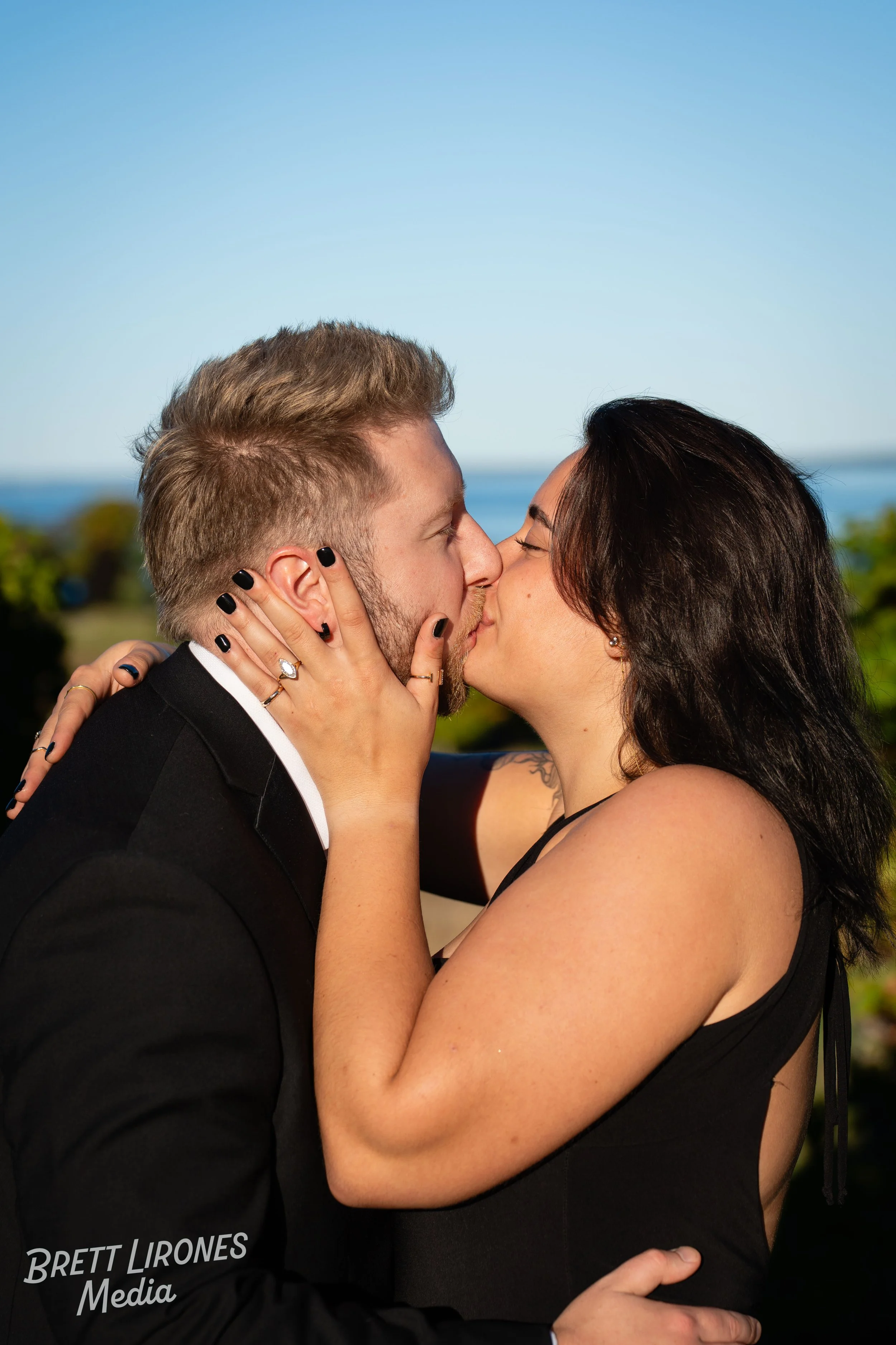 A couple kissing outdoors with a scenic background of trees and a body of water.