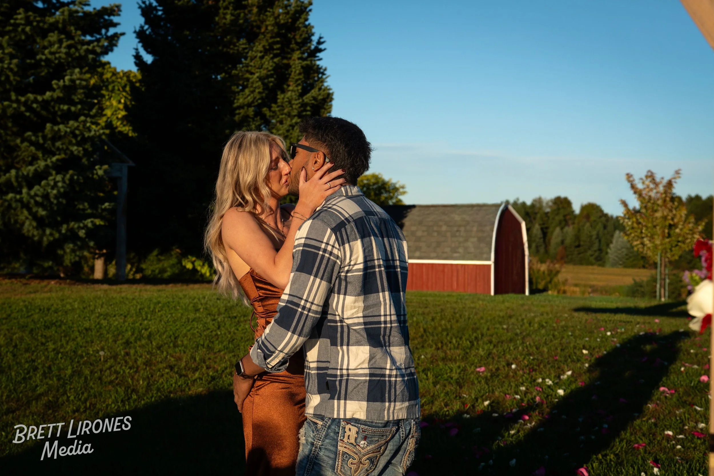 A couple sharing a kiss outdoors in a rural setting with a red barn and green trees in the background during sunset.
