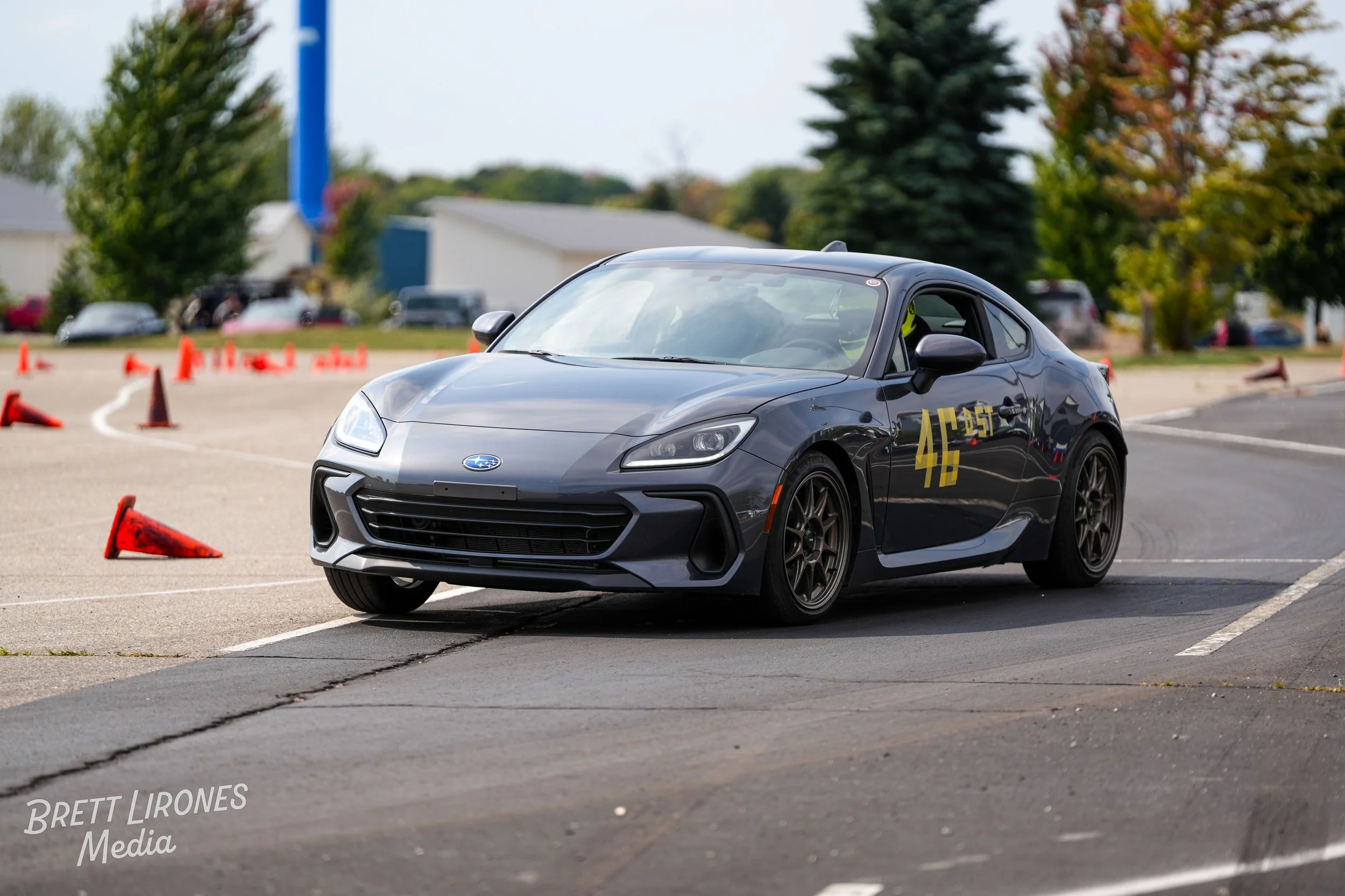 A dark gray Subaru sports car navigating through a parking lot marked with orange cones during a driving skills course. The car has the number 45 and the word 'BLAST' written on the side. The background includes trees and other parked cars.