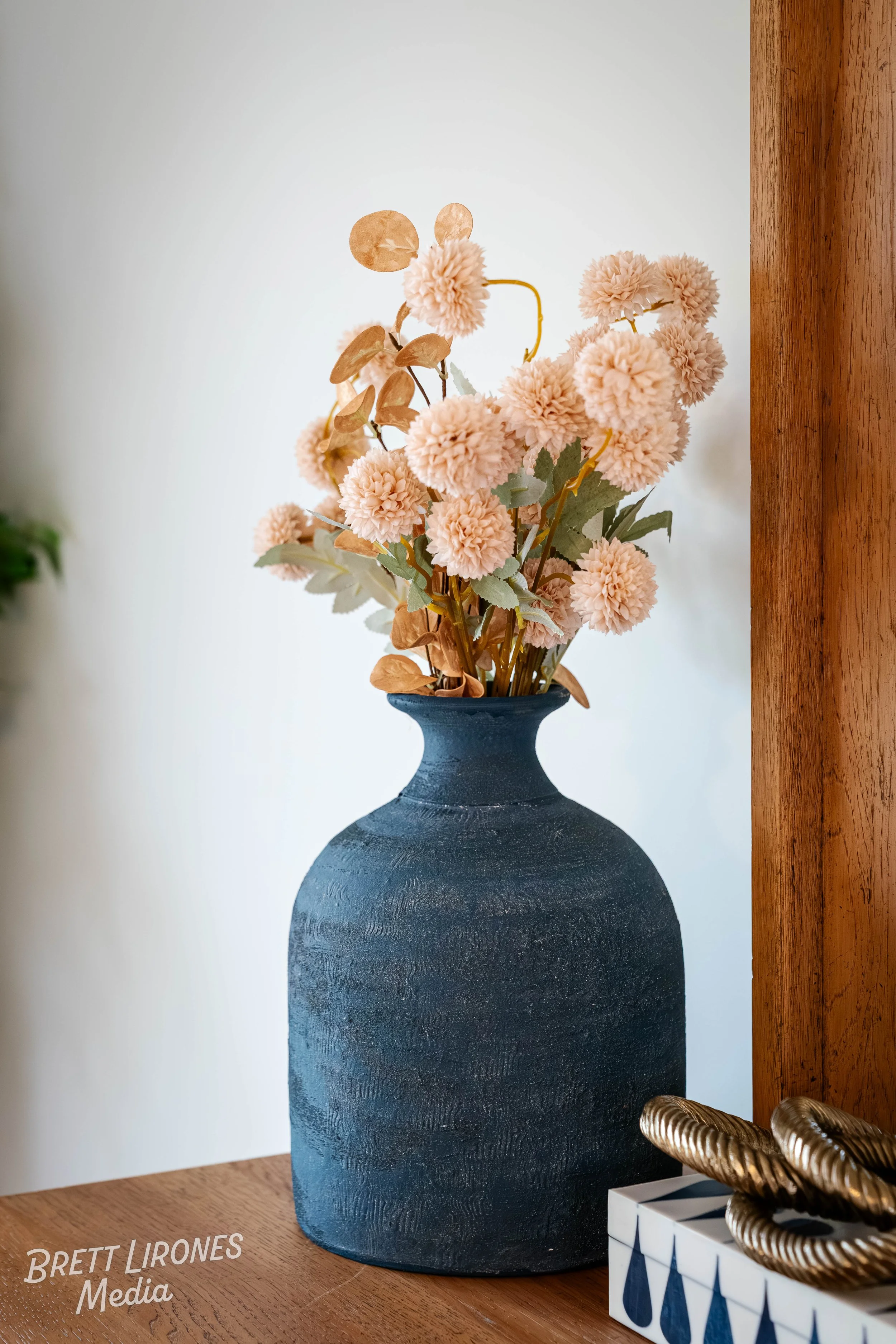 A ceramic vase with a textured dark blue finish containing beige pom-pom flowers and dried leaves, next to a white box with a blue pattern and two gold-colored coiled decorative objects, on a wooden surface against a light-colored wall.