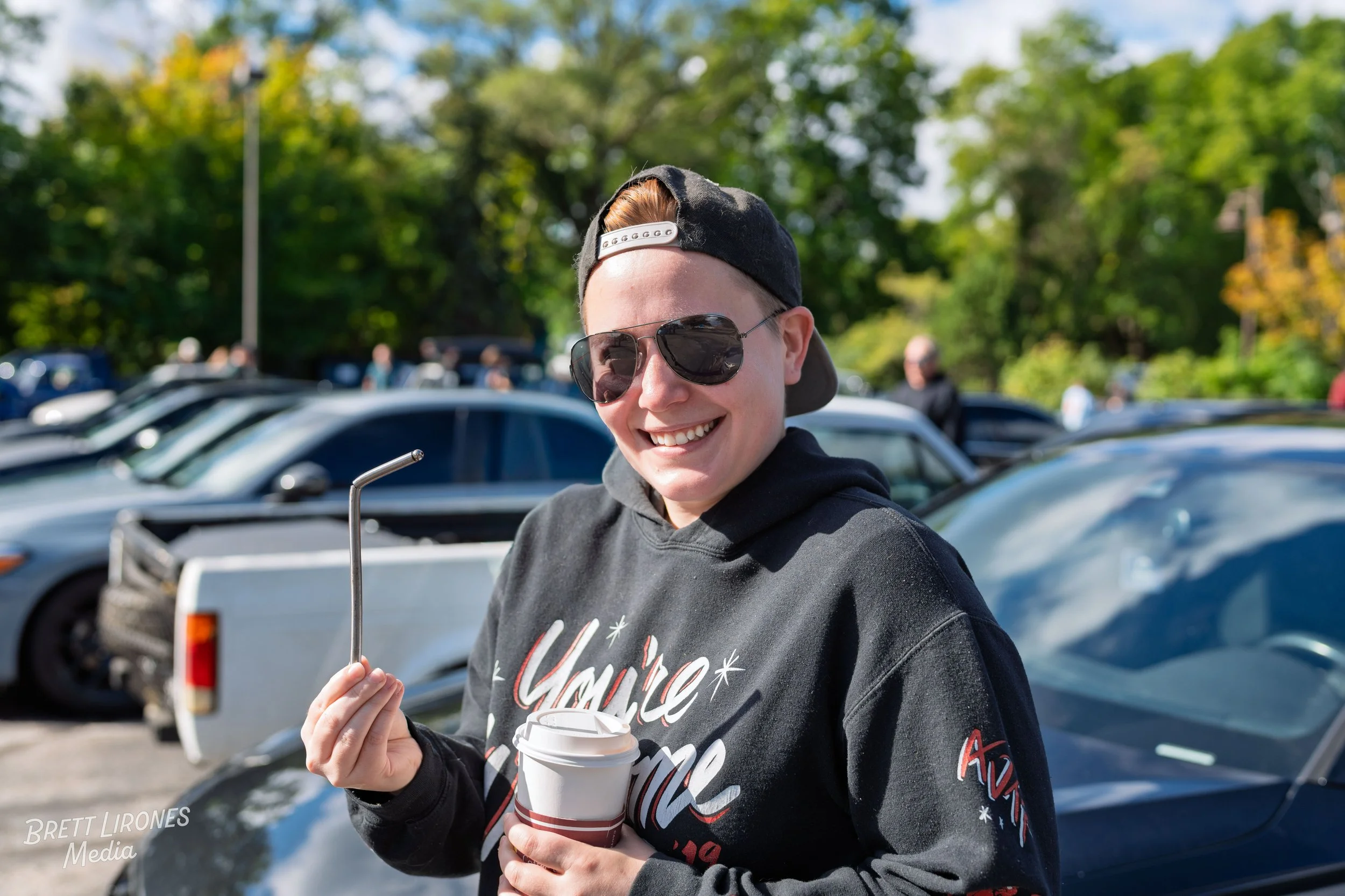 Smiling person wearing sunglasses, black hoodie, and backwards cap, holding a disposable coffee cup and a metal straw in a parking lot surrounded by cars and green trees.