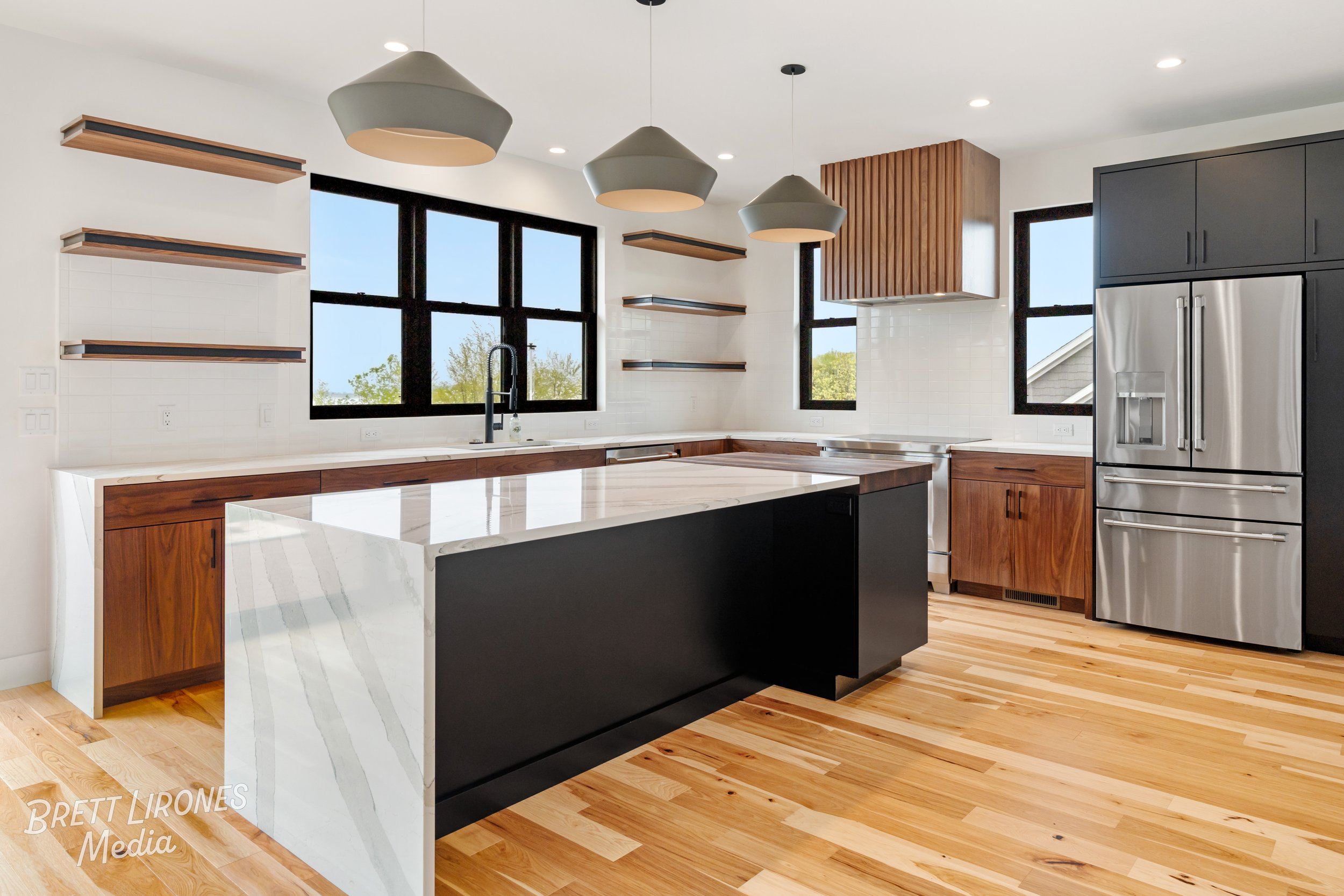 Modern kitchen with black window frames, wooden cabinets, stainless steel refrigerator, and a large island with a marble top. Open shelving on the wall, wood floors, and pendant lights.