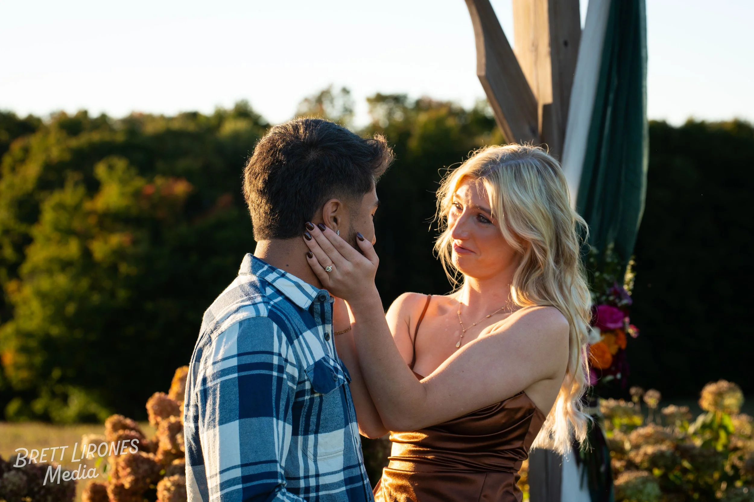 A woman with blonde hair holding a man's face with her hands, standing outdoors during sunset, with a decorative arch and greenery in the background.