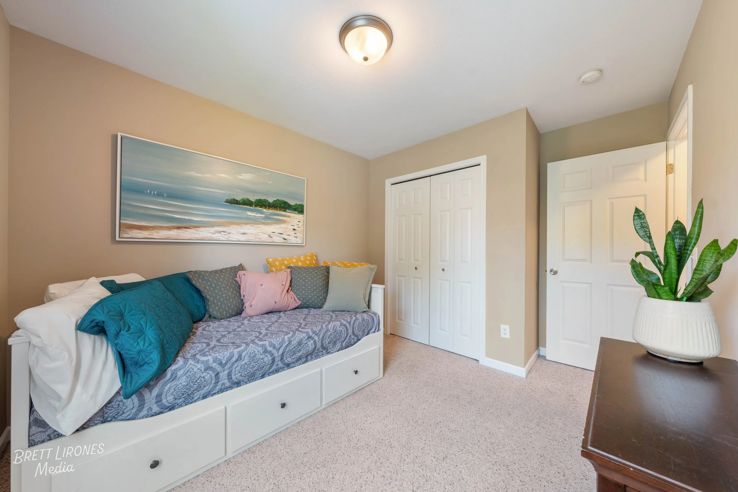 A bedroom with a beige wall, white closet doors, and a white daybed with blue, gray, pink, and yellow pillows, a painting of a beach scene above the bed, a dark wooden dresser with a pot of green houseplants, and beige carpeted flooring.