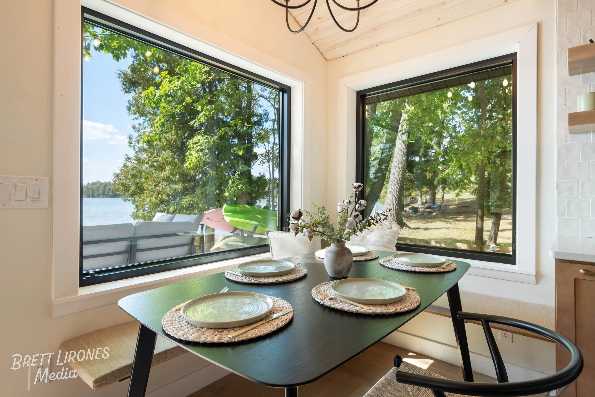 Dining table set with four placemats, plates, and cutlery, next to large windows overlooking trees and a river outside, with a vase of flowers in the center.